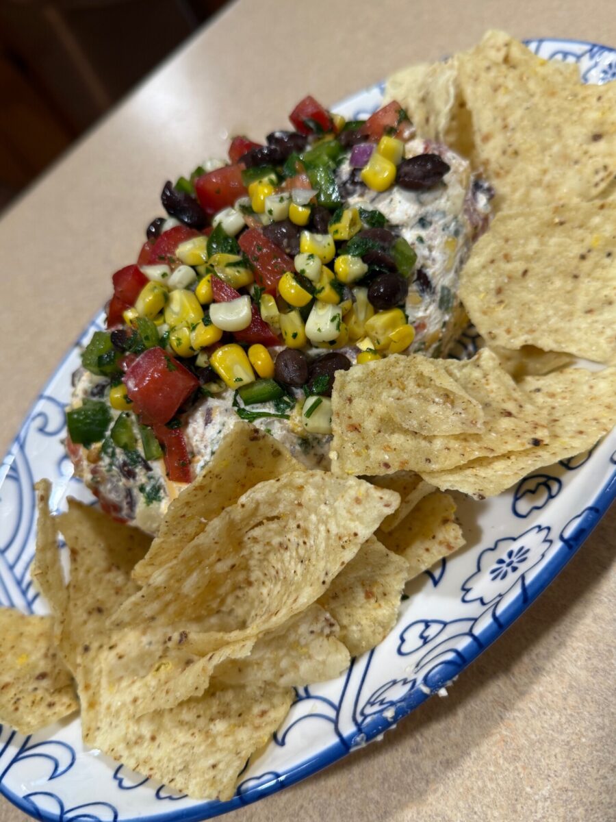 A plate of tortilla chips surrounds a mound of dip topped with corn, black beans, tomatoes, and chopped green herbs on a decorative blue and white plate.