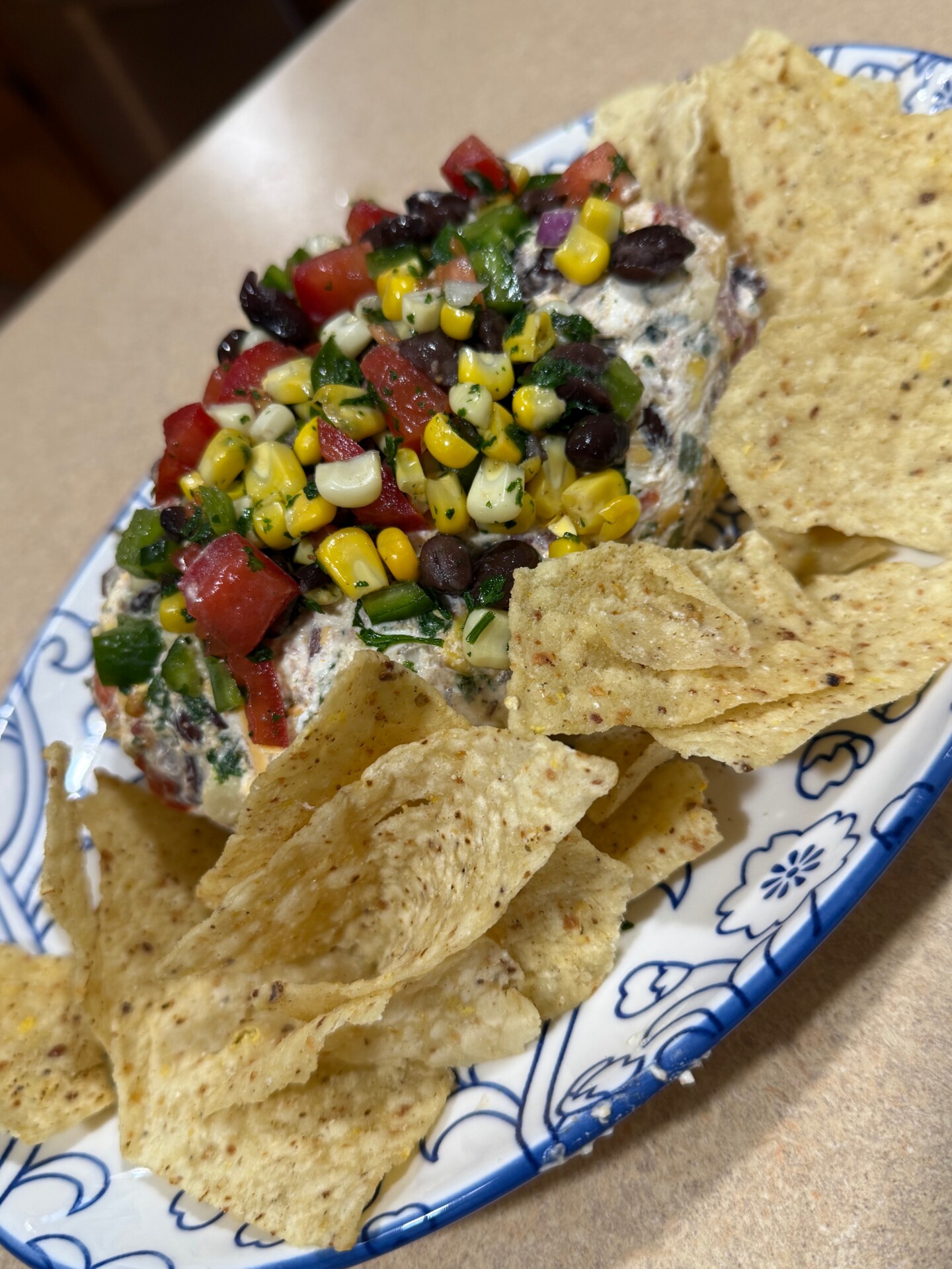 A platter with tortilla chips surrounds a mound of cream cheese dip topped with colorful black beans, corn, tomatoes, green onions, and herbs on a blue and white plate.