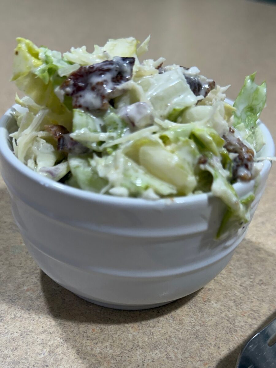 A white bowl filled with creamy coleslaw that includes chopped lettuce, cabbage, dark raisins, and diced vegetables, sitting on a light brown countertop.