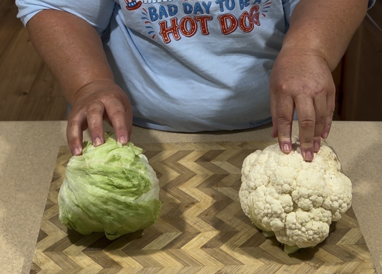 A person wearing a light blue shirt with a hot dog graphic holds a head of lettuce with their left hand and a head of cauliflower with their right hand on a wooden cutting board.