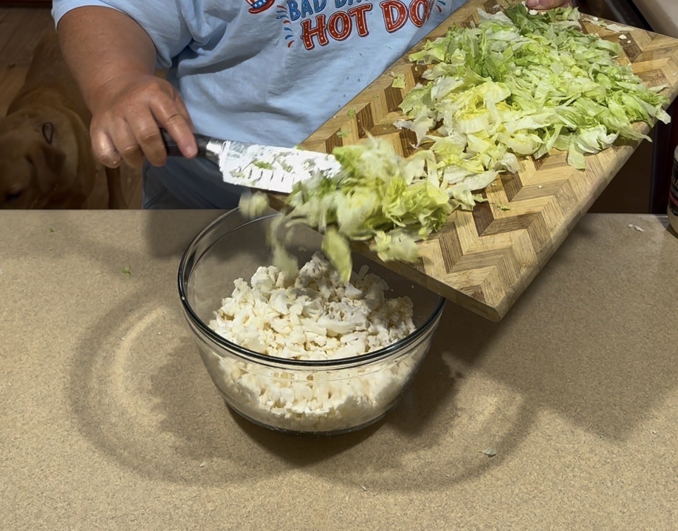 A person adds chopped lettuce from a wooden cutting board into a glass bowl filled with diced cauliflower on a kitchen counter. A dog is partially visible in the background.