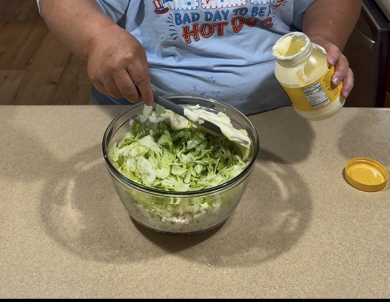 A person wearing a blue Bad Day to Be a Hot Dog shirt is adding mayonnaise from a jar into a glass bowl filled with chopped lettuce on a kitchen counter.