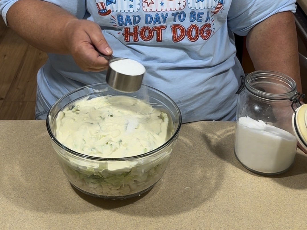 A person in a “Bad Day to Be a Hot Dog” shirt holds a measuring cup of sugar over a bowl filled with layered ingredients, next to an open jar of sugar on a kitchen counter.