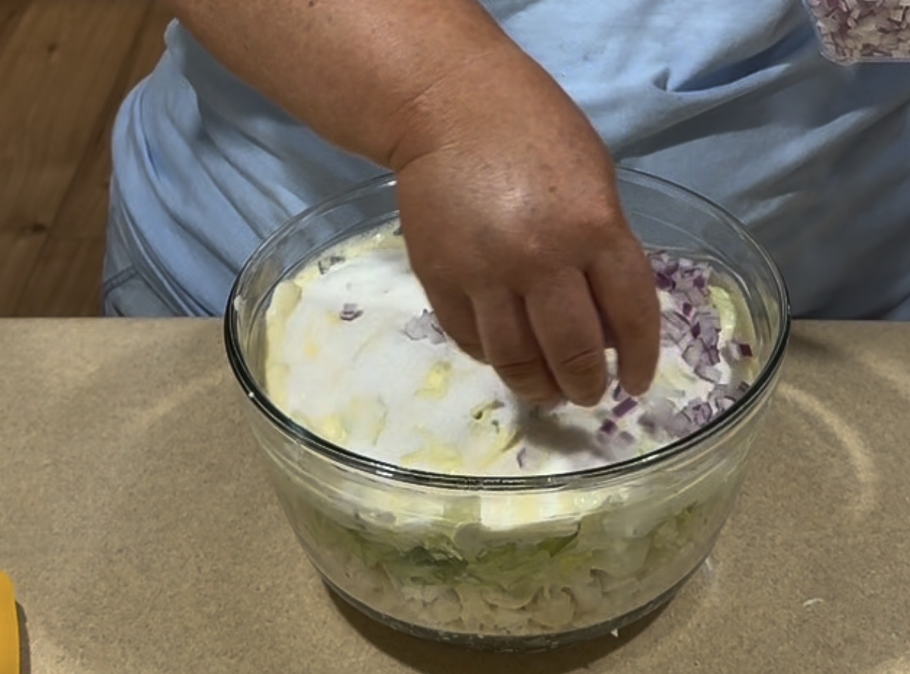 A person sprinkles chopped red onion onto a layered salad in a glass bowl, with visible layers of lettuce, mayonnaise, and other ingredients.
