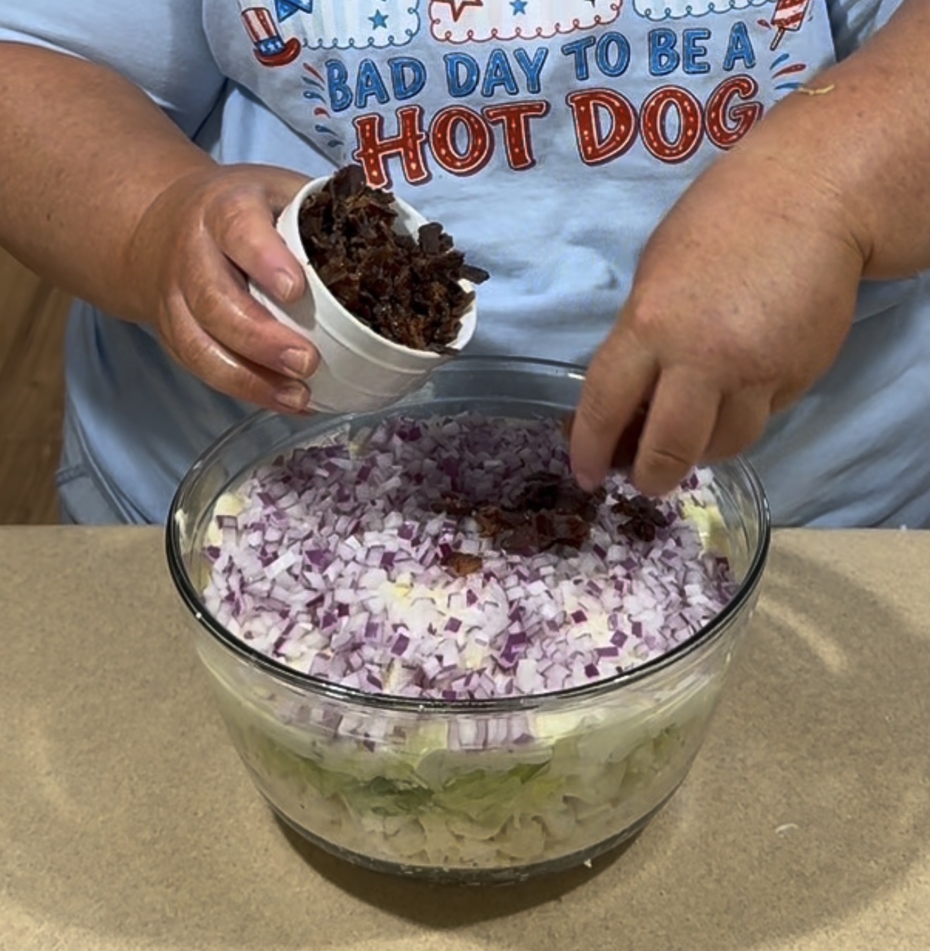A person wearing a “Bad Day To Be A Hot Dog” shirt sprinkles chopped bacon from a cup onto a layered salad in a glass bowl, with chopped red onions visible on top.
