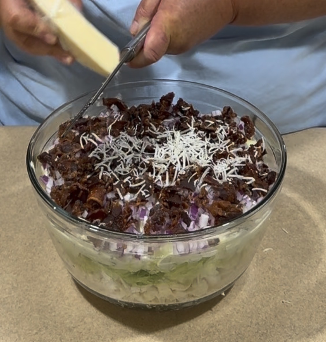 A person grates cheese over a glass bowl filled with a layered salad, including lettuce, chopped red onions, bacon, and shredded cheese, on a kitchen counter.