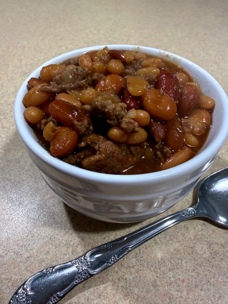 A white bowl filled with chili containing beans, carrots, and ground meat, sits on a tan countertop next to a silver spoon.