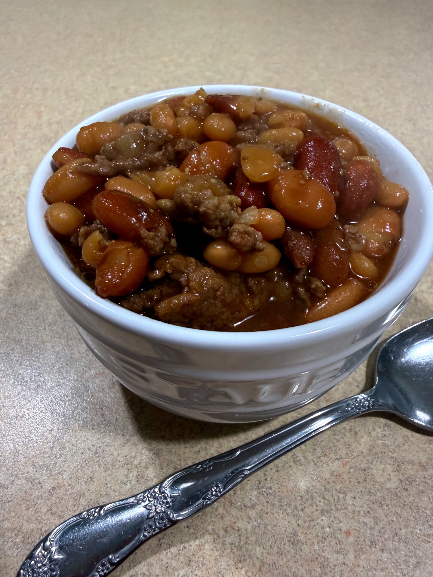 A white bowl filled with chili containing beans, carrots, and ground meat, sits on a tan countertop next to a silver spoon.