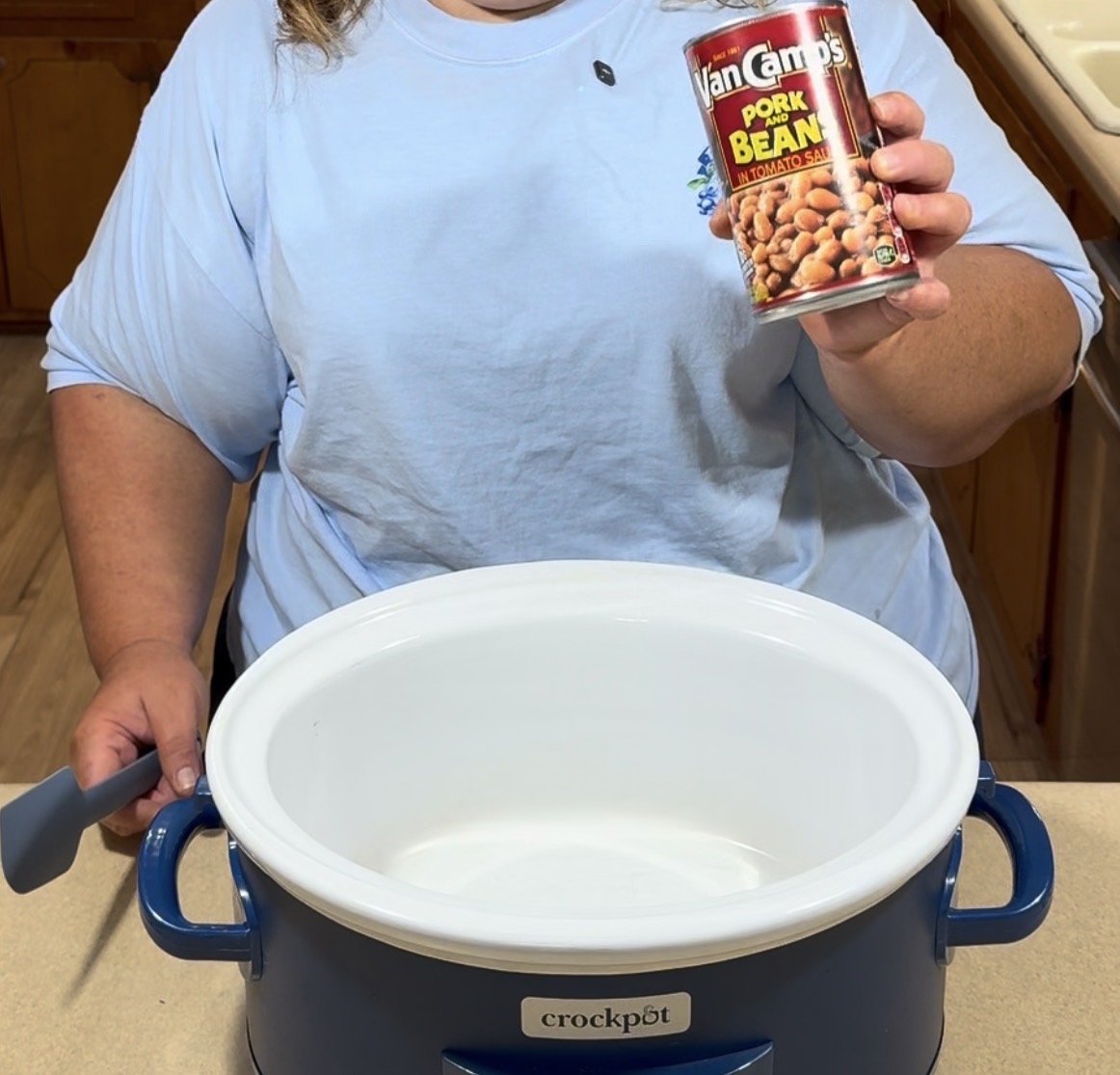A person holding a can of Van Camp’s Pork and Beans above an empty blue crockpot, preparing to add the beans. The person is also holding a spatula in the other hand.