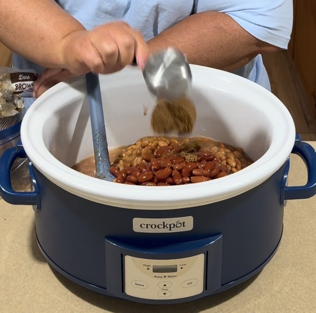 A person adds seasoning to a crockpot filled with mixed beans using a measuring cup, while stirring with a blue spatula. The crockpot is blue and sits on a kitchen counter.
