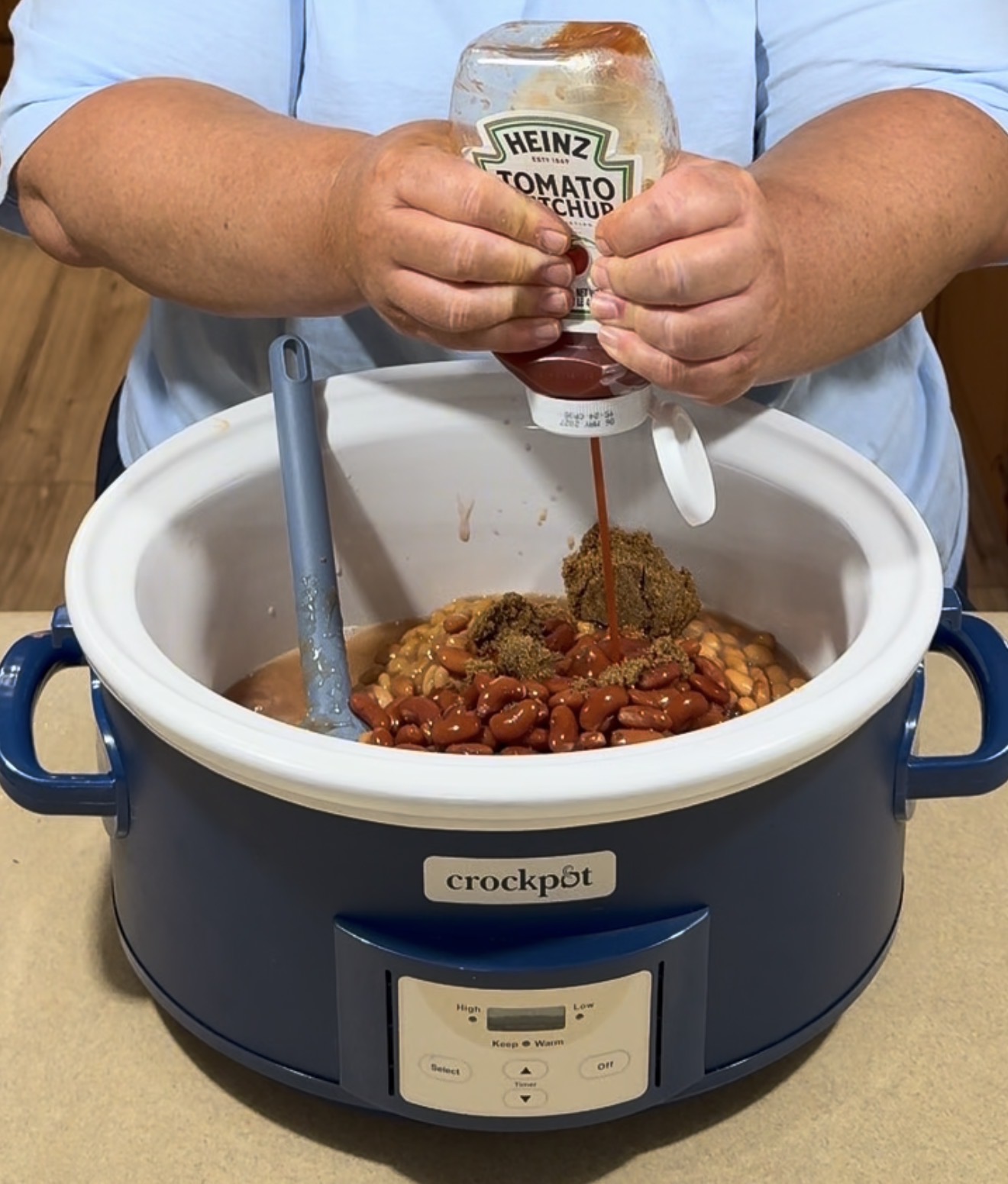 A person squeezes Heinz tomato ketchup into a crockpot filled with beans and brown sugar, with a blue spatula in the pot. The crockpot is blue and placed on a kitchen counter.