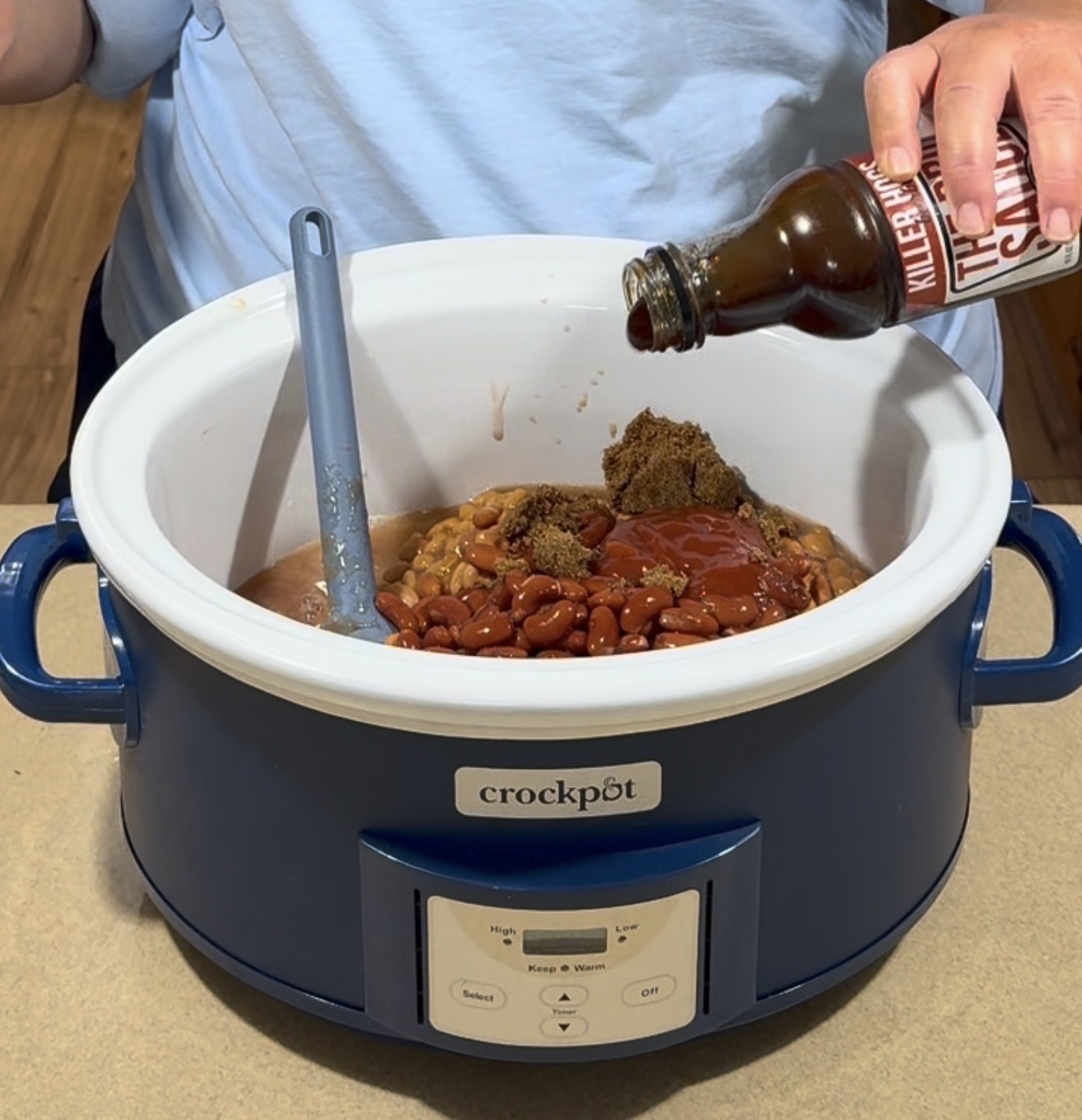 A person pours barbecue sauce into a blue Crockpot filled with beans, brown sugar, ketchup, and other ingredients, preparing to make a dish. A blue spatula rests in the mixture.
