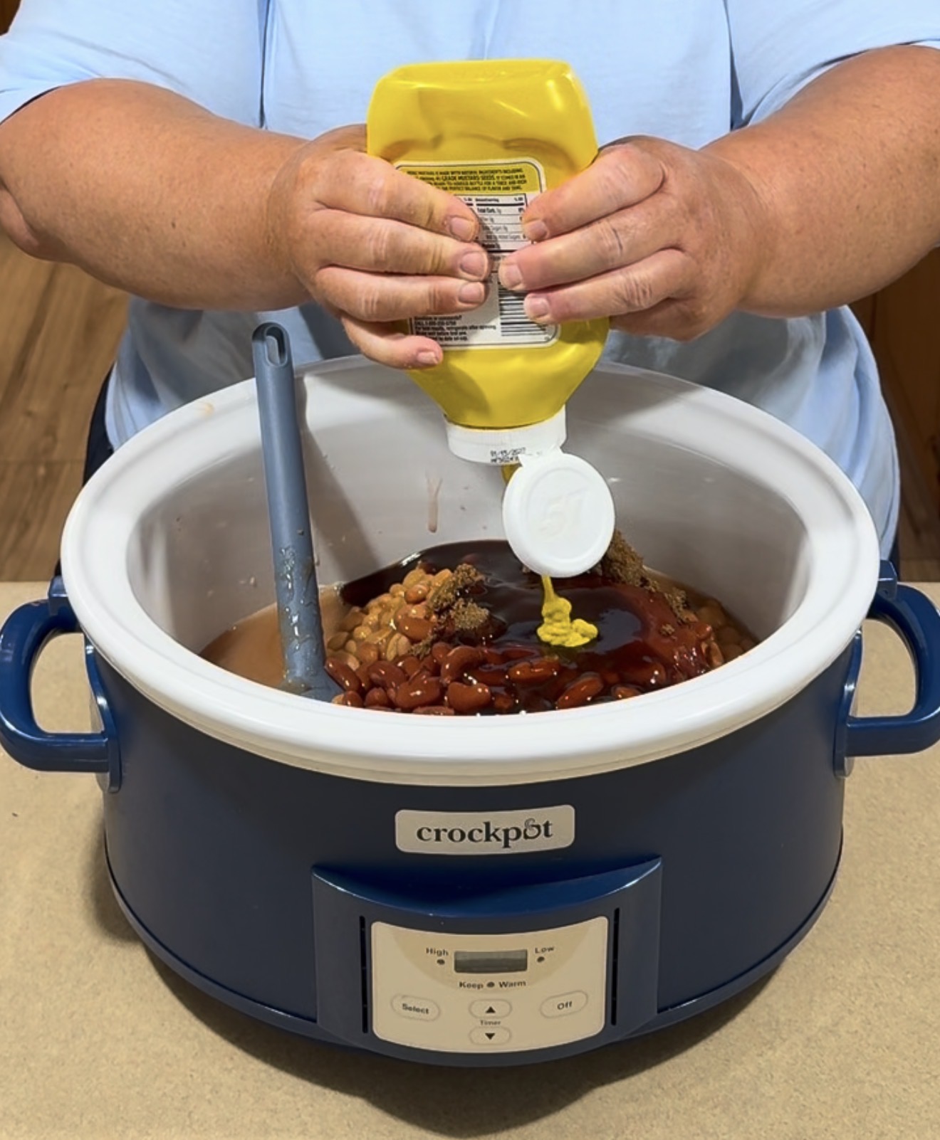 A person squeezes yellow mustard into a blue Crock-Pot filled with beans and sauce, mixing ingredients with a spatula on a kitchen counter.