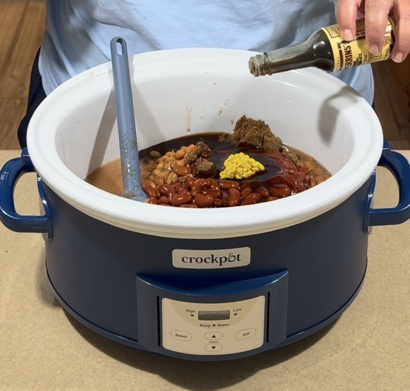 A person pours Worcestershire sauce into a blue Crock-Pot filled with beans, ketchup, mustard, and spices, using a spatula to mix the ingredients.