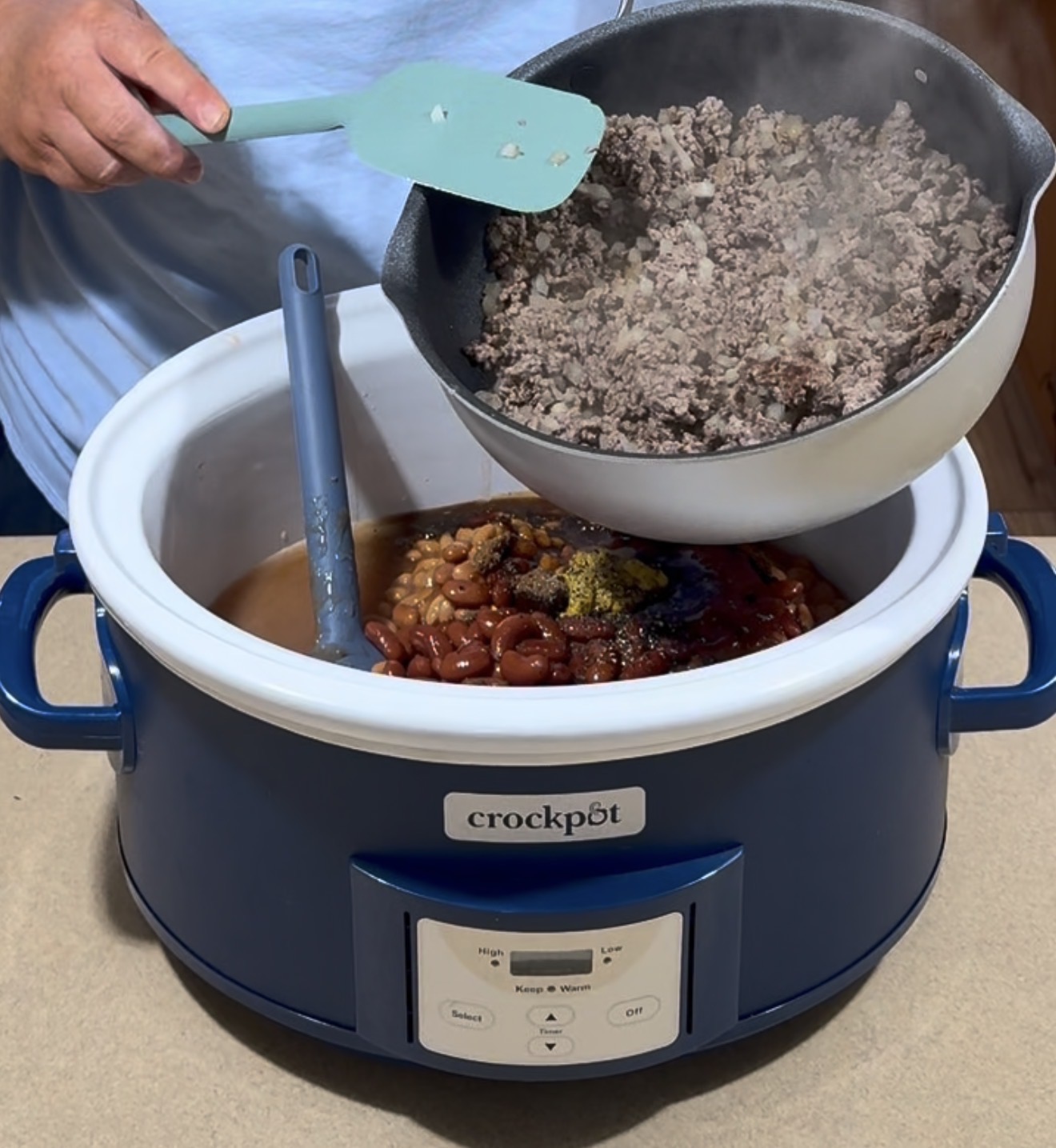 A person pours cooked ground beef from a skillet into a Crockpot filled with beans and spices, preparing a slow-cooked meal.