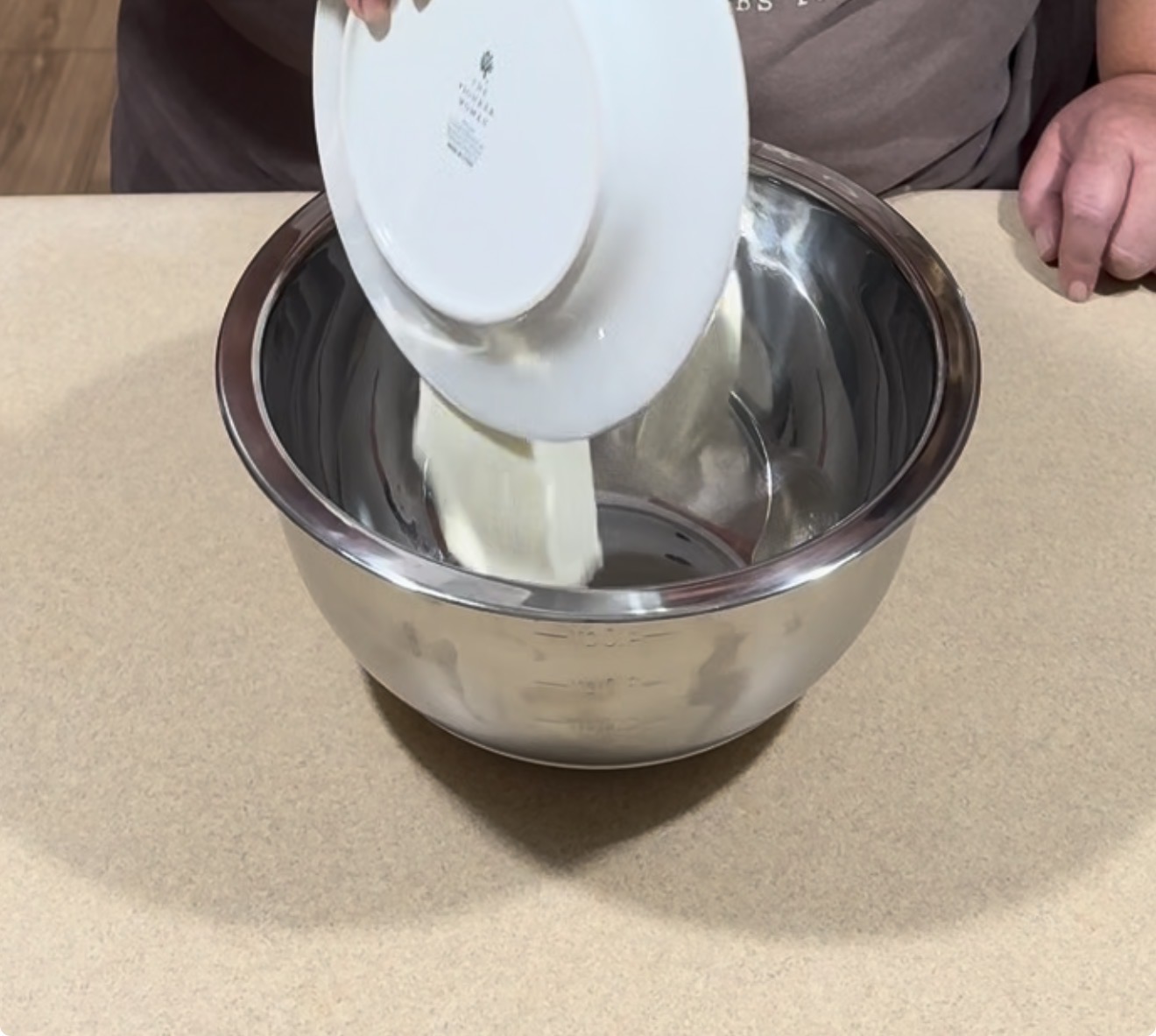 Person pouring flour from a white plate into a large metal mixing bowl on a beige countertop.