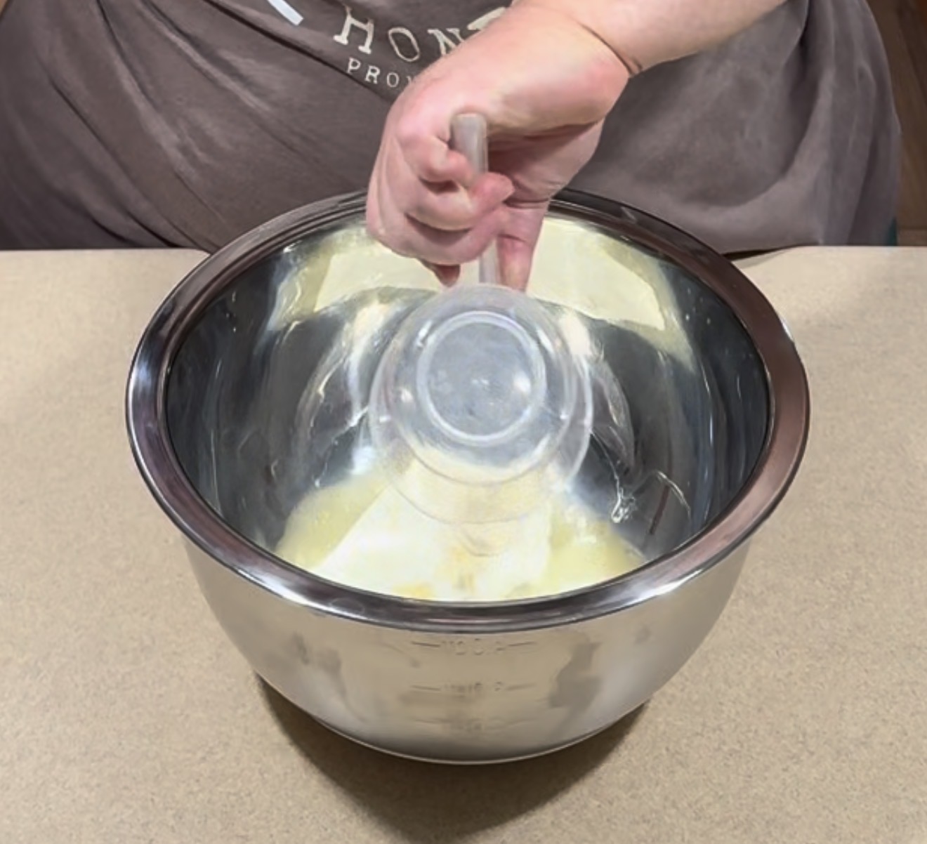 A person holds a glass bowl, pouring its contents into a large metal mixing bowl with a light yellow mixture inside, on a beige countertop.
