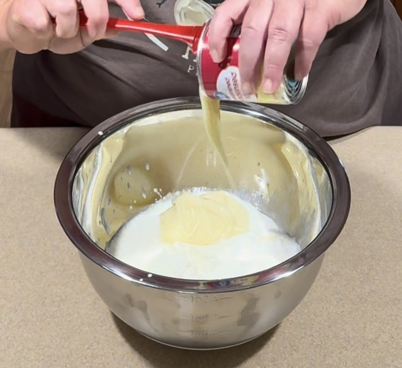 Hands pour condensed milk from a can into a metal mixing bowl already containing a white liquid, possibly cream or milk, using a red spatula. The bowl is placed on a beige countertop.