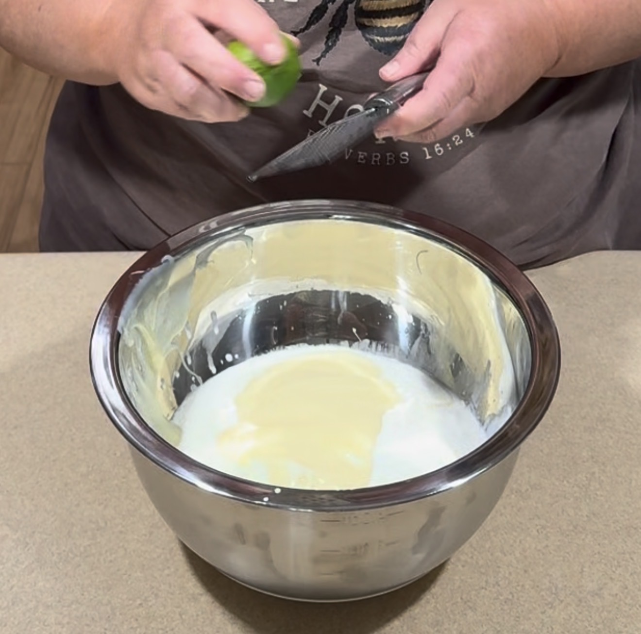 A person grates a lime over a large metal mixing bowl filled with a creamy mixture on a kitchen counter.