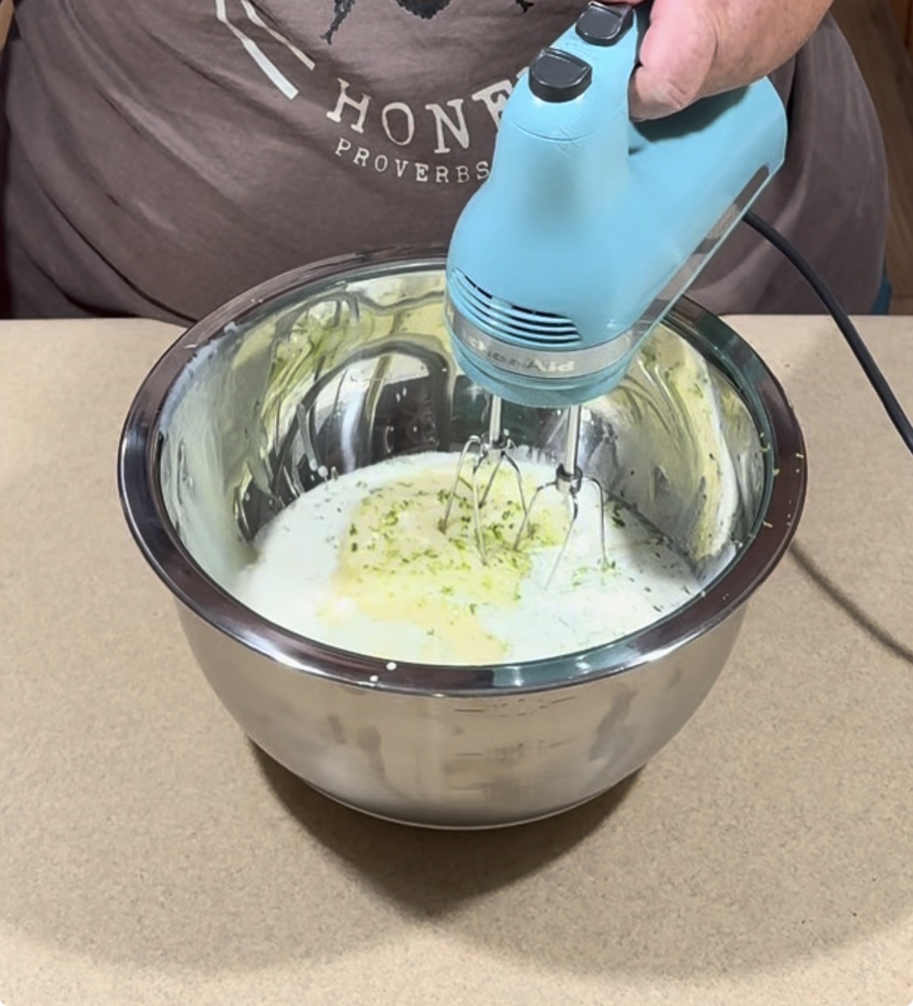 A person uses a blue hand mixer to blend ingredients in a large metal bowl on a countertop. The mixture appears creamy with green zest on top.