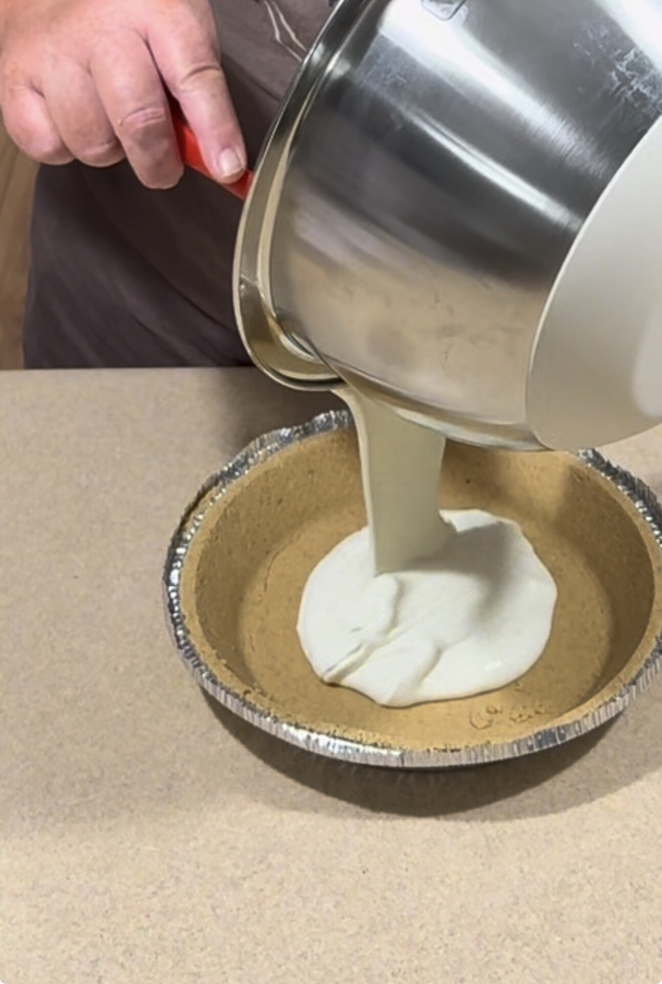 A person pours creamy pie filling from a mixing bowl into a prepared graham cracker crust on a kitchen counter.