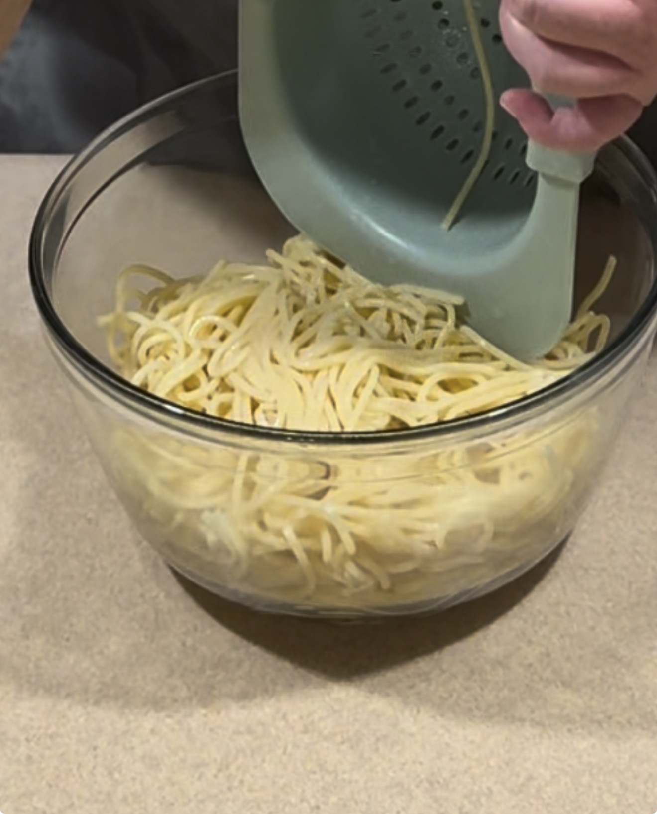A person pours cooked spaghetti from a green colander into a large glass bowl on a beige countertop.
