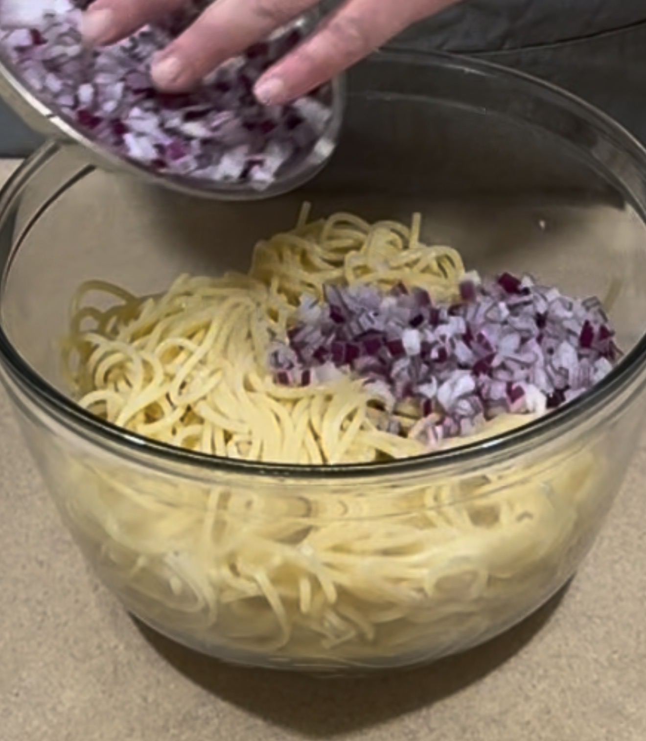 A hand adds chopped red onions from a small bowl into a larger glass bowl filled with cooked spaghetti noodles on a countertop.
