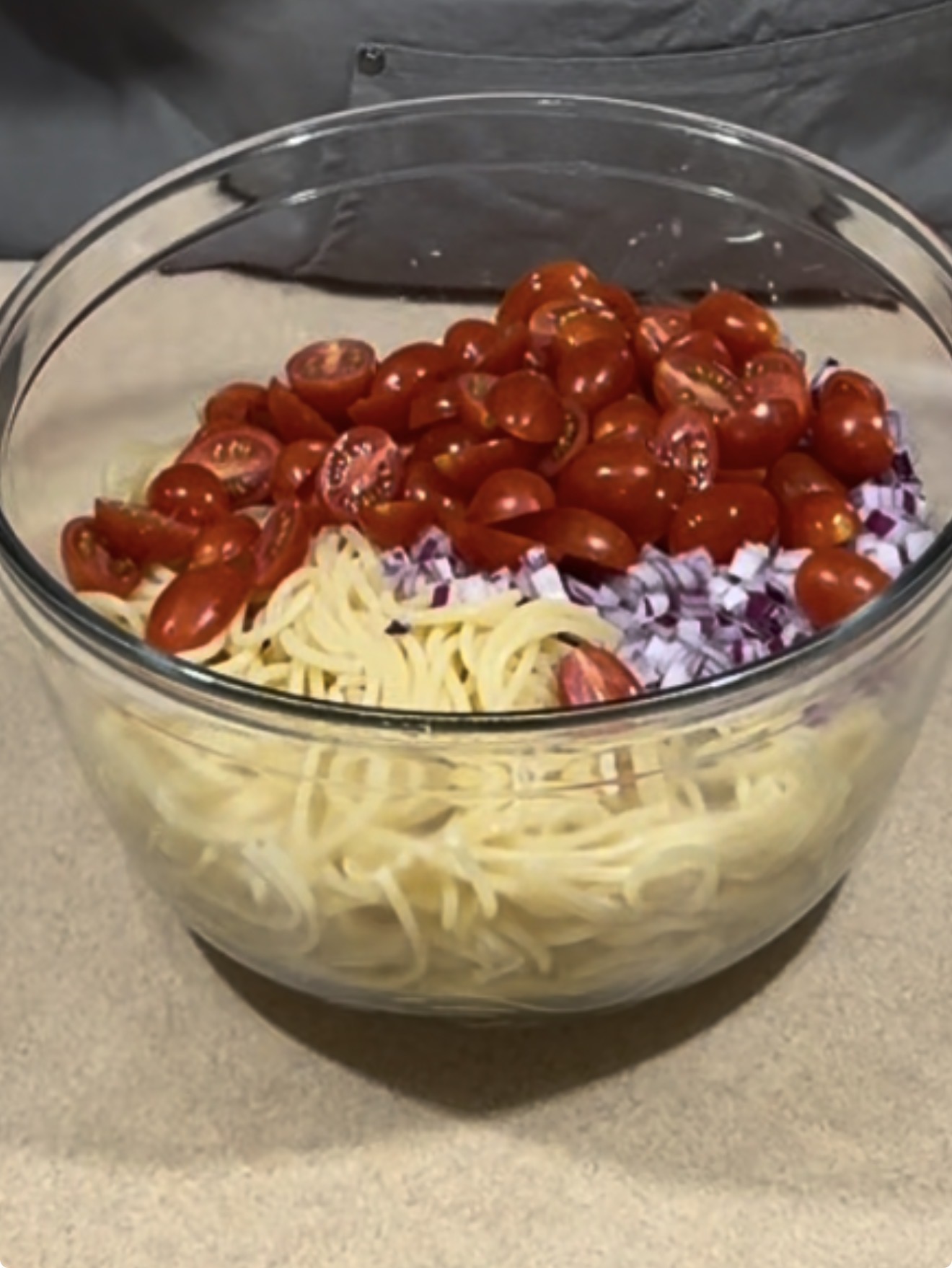 A glass bowl filled with cooked spaghetti, halved cherry tomatoes, and chopped red onions sits on a beige countertop.