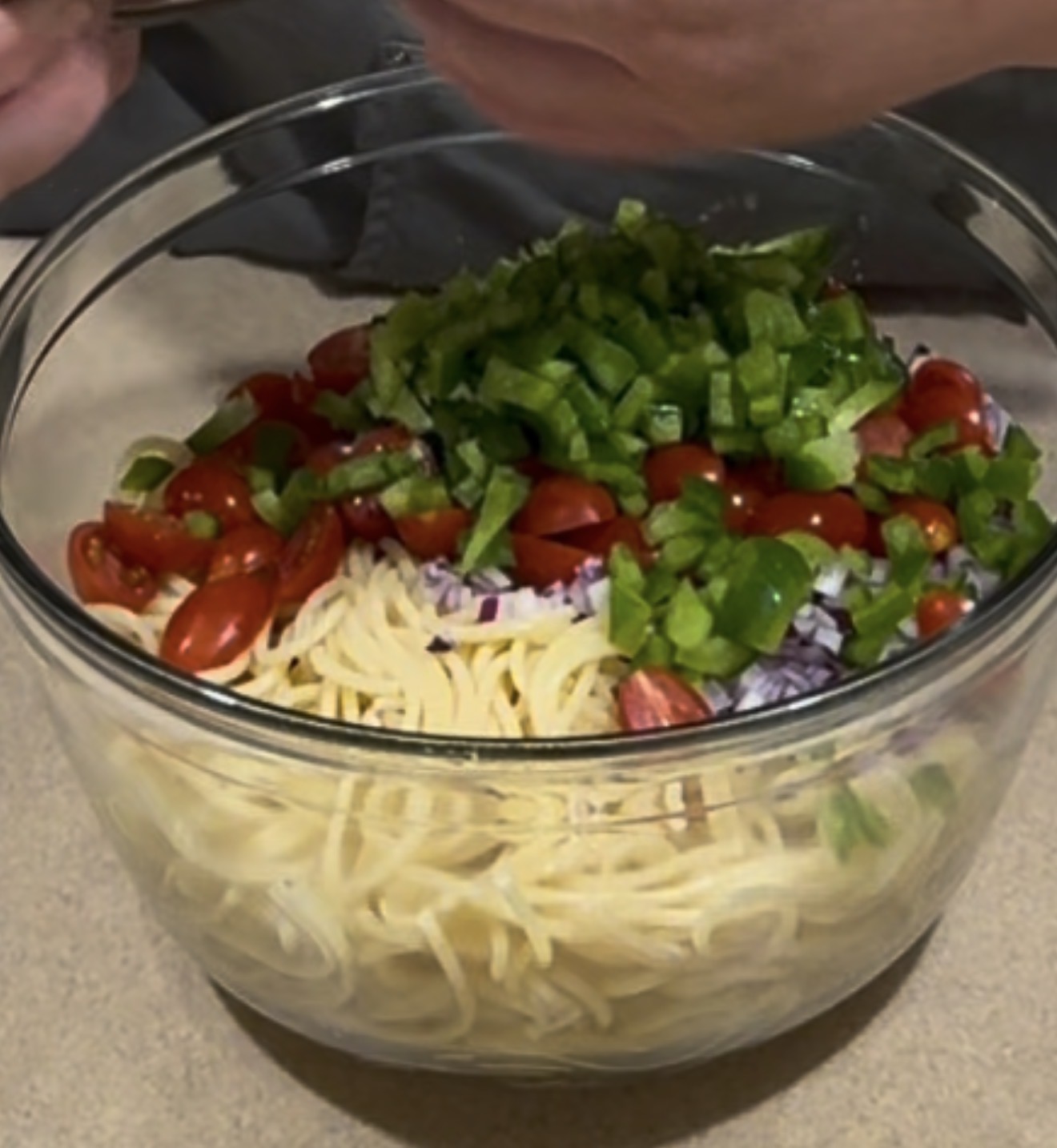 A glass bowl filled with cooked spaghetti, chopped green bell peppers, grape tomatoes, and diced red onions on a countertop.