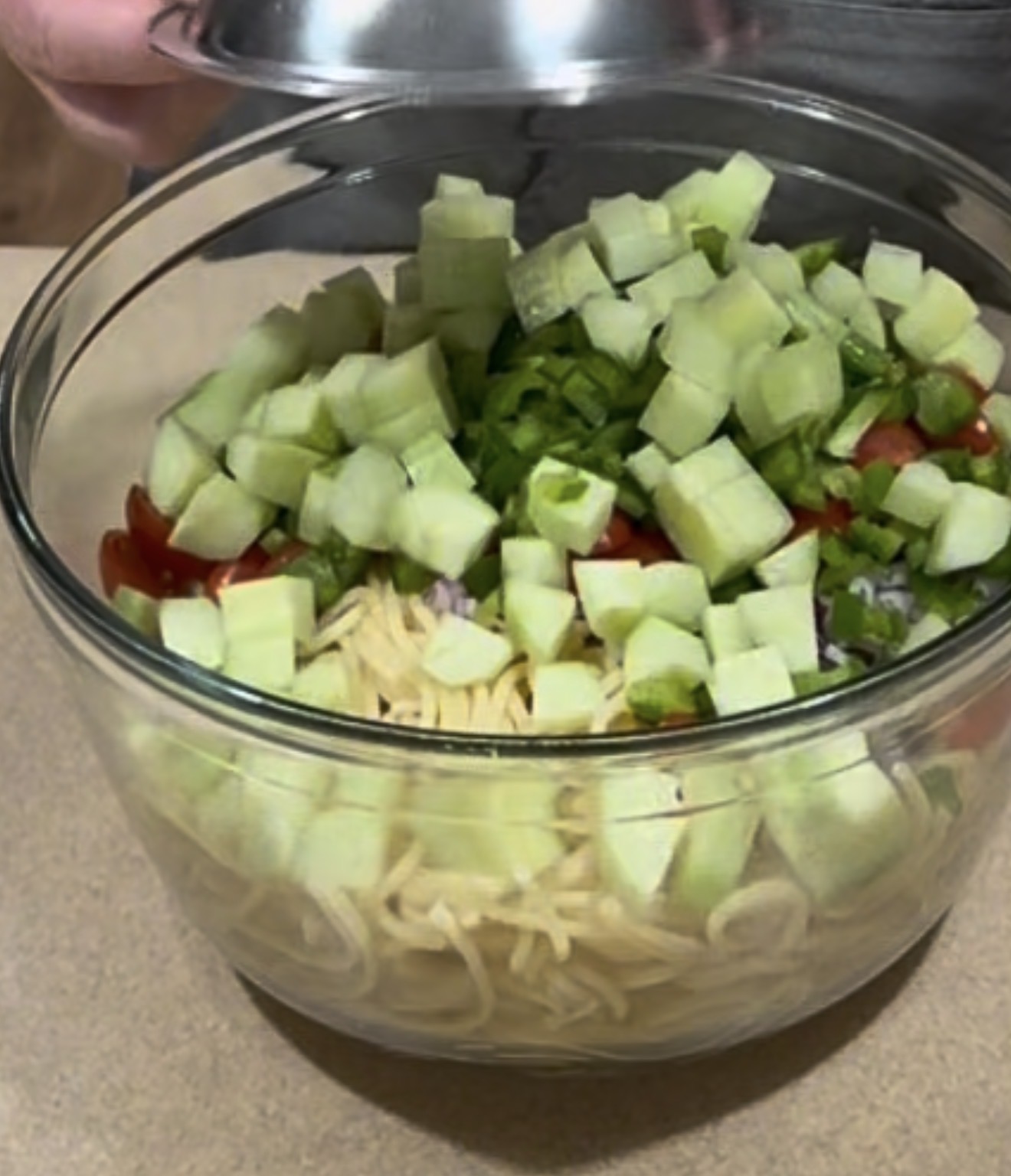 A glass bowl filled with uncooked spaghetti noodles, diced cucumbers, chopped green peppers, tomatoes, and onions sits on a countertop. A hand lifts a metal lid off the bowl.
