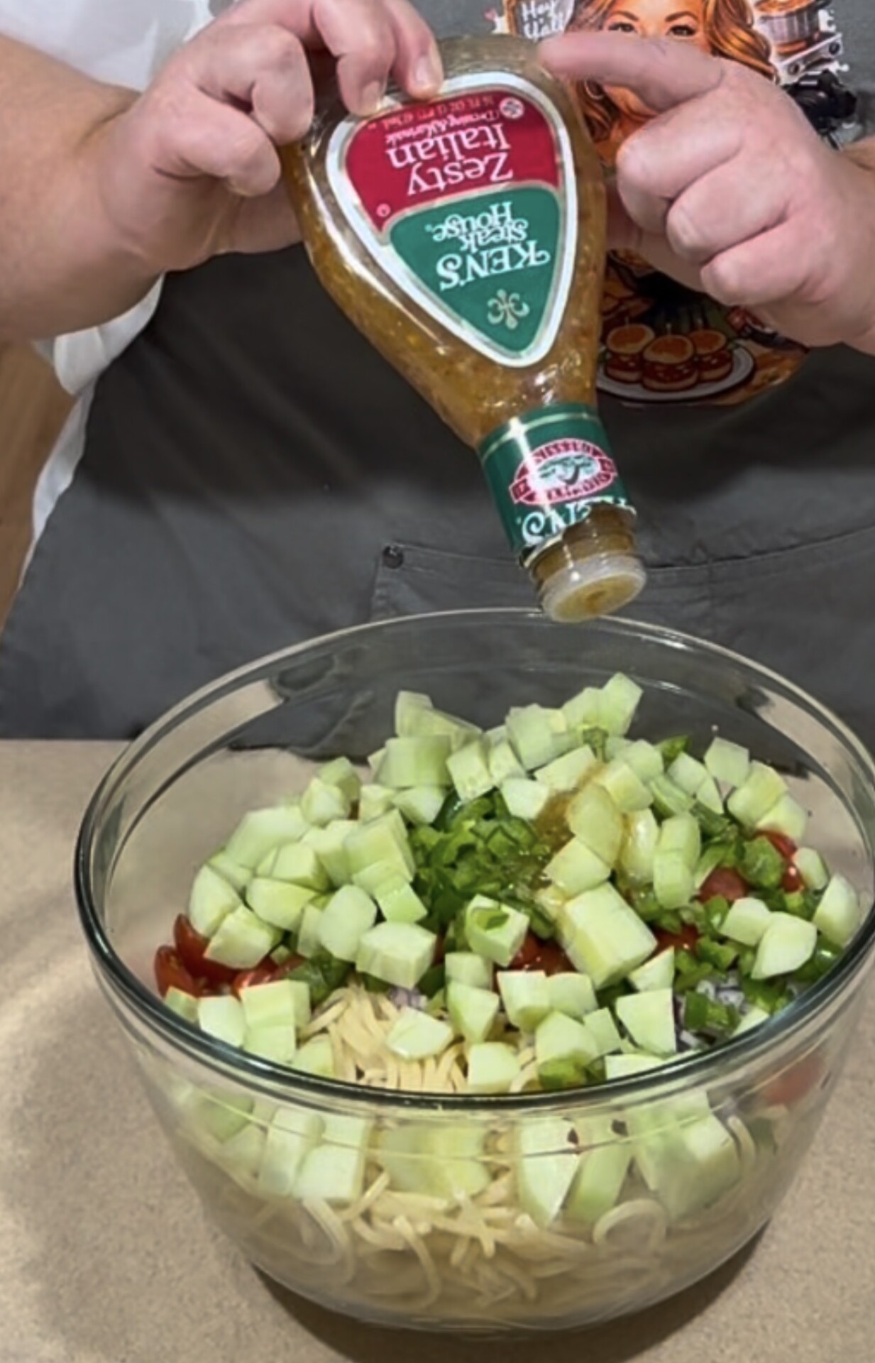 A person pours Italian dressing onto a salad with cucumbers, tomatoes, and pasta in a glass bowl on a countertop.