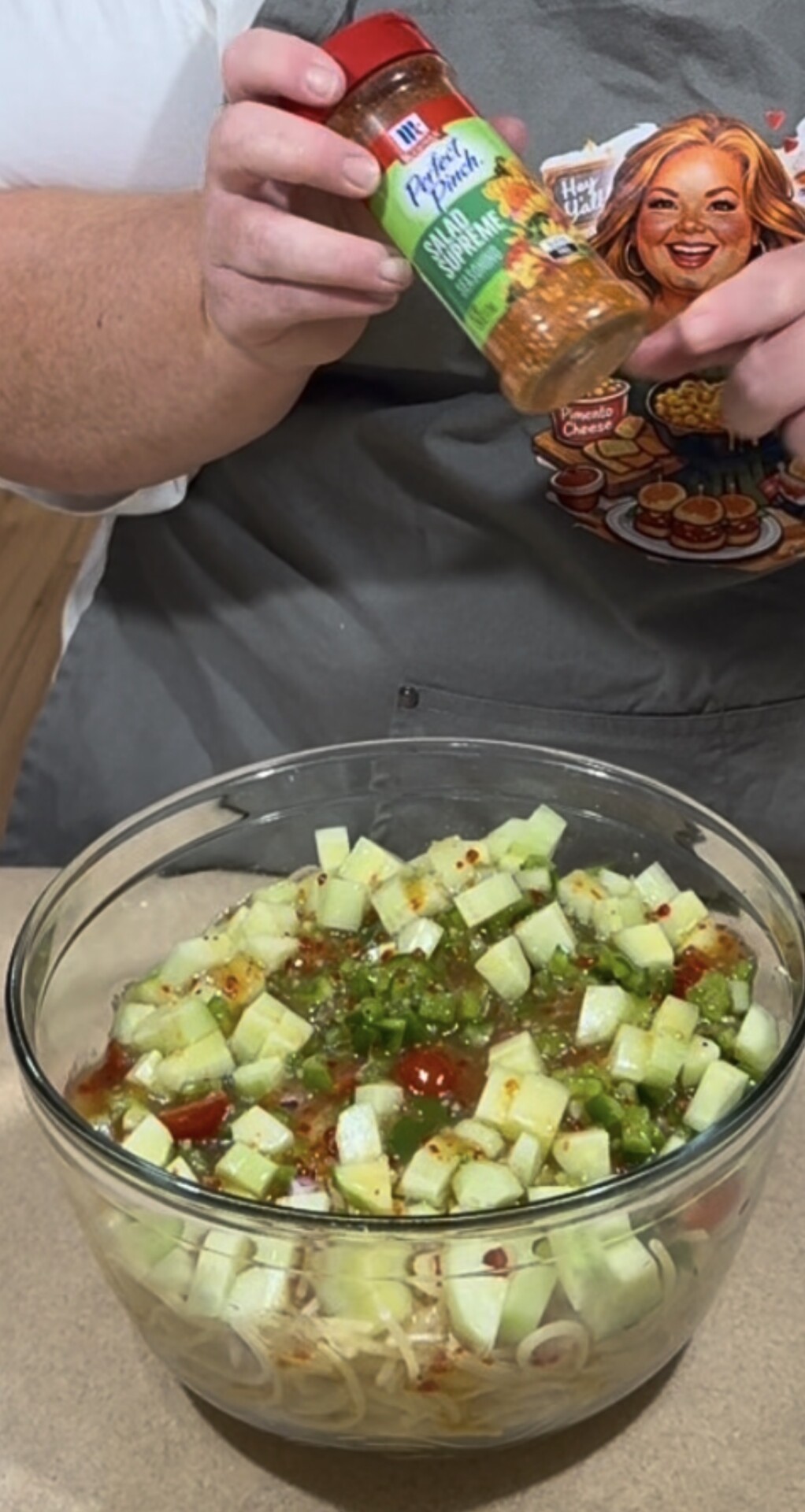 A person holds a spice bottle above a glass bowl filled with chopped cucumbers, vegetables, and dressing. The person is wearing a gray apron with a cartoon image and food illustrations on it.
