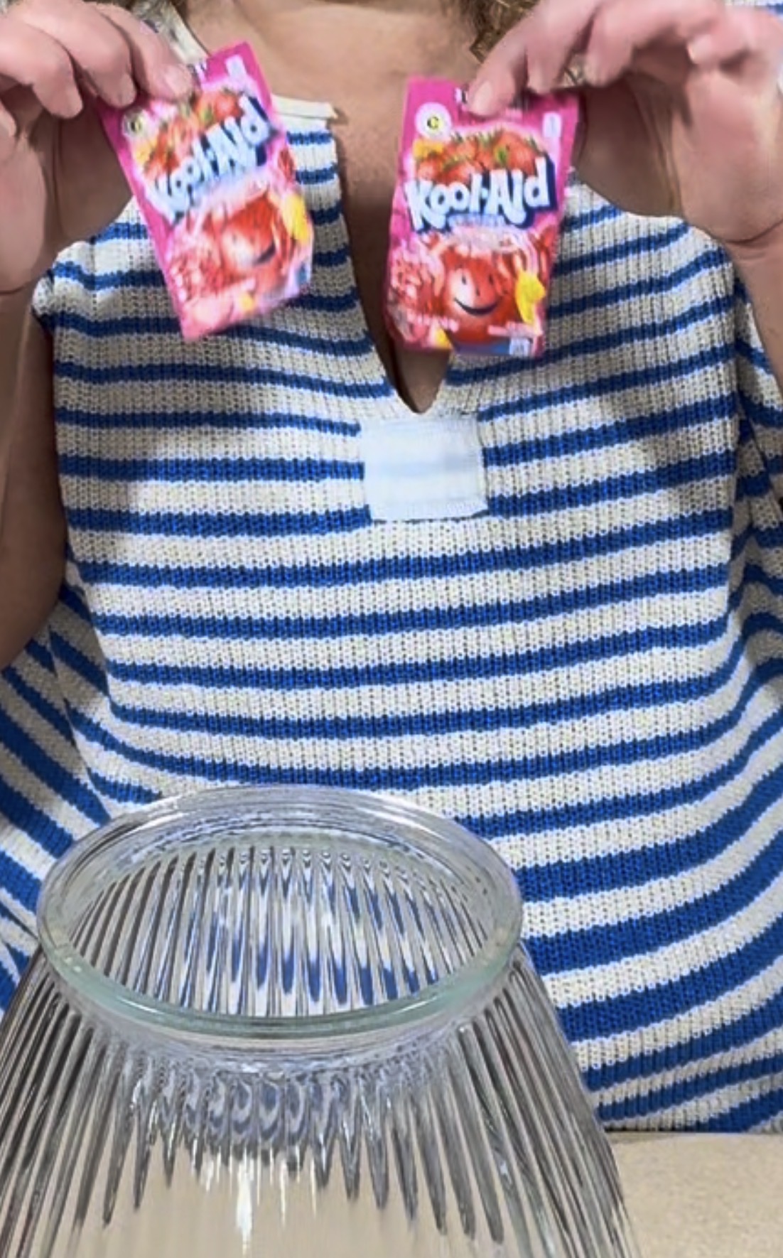 A person in a blue and white striped shirt holds two packets of cherry Kool-Aid above a large empty glass bowl.