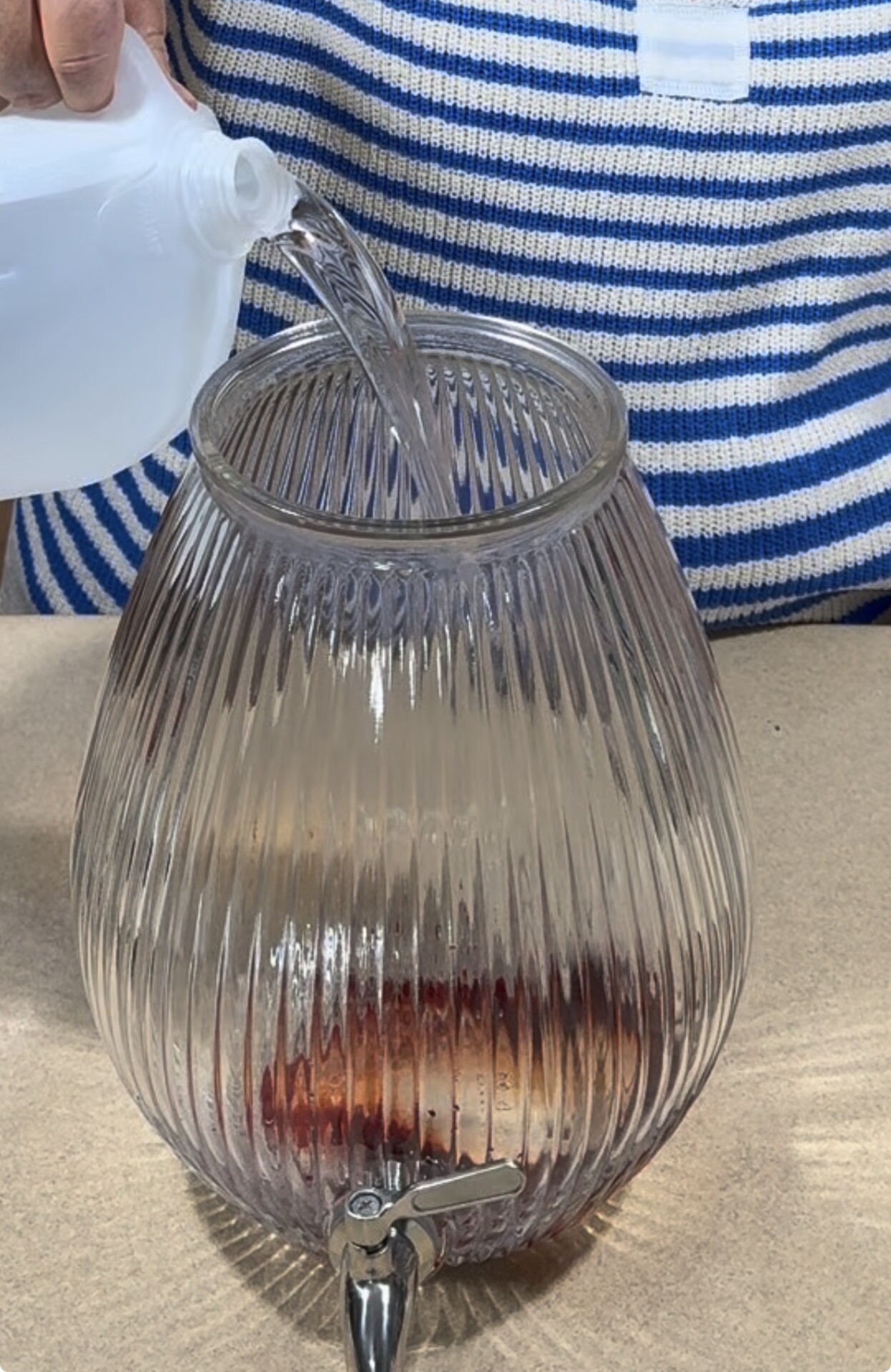 A person pours liquid from a white bottle into a large, ribbed glass dispenser with a spigot, which contains a small amount of dark liquid at the bottom.