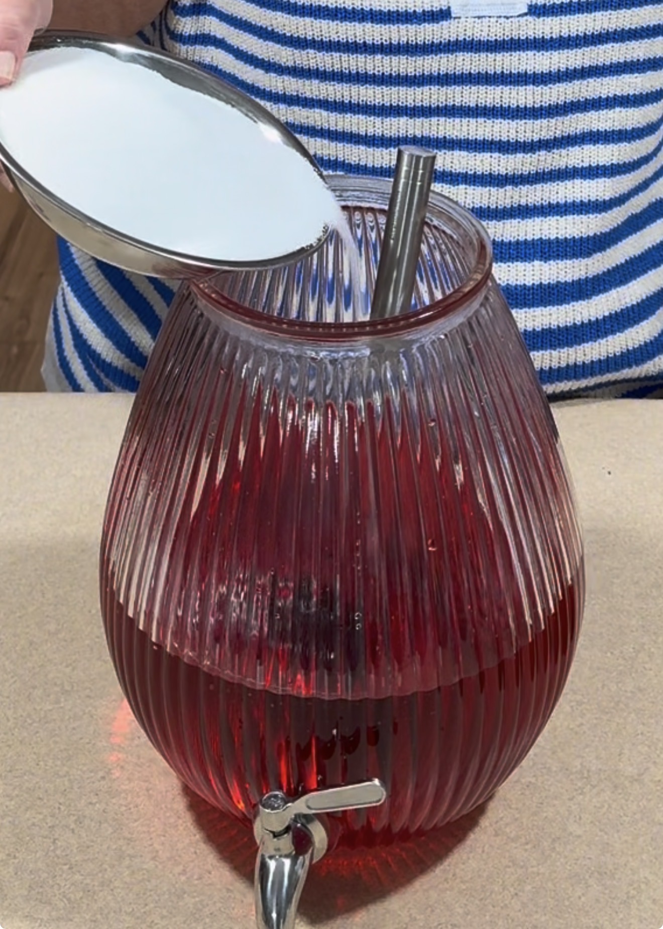 A person pours a large scoop of white sugar into a glass dispenser filled with red liquid, possibly punch, using a metal straw for stirring. The person is wearing a blue and white striped shirt.