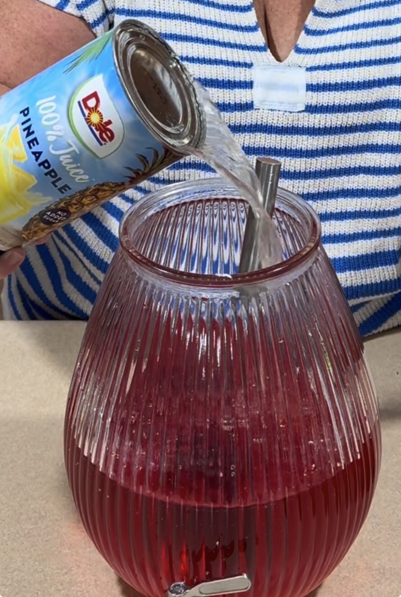 A person in a striped shirt pours Dole pineapple juice from a can into a large glass beverage dispenser filled with red liquid.
