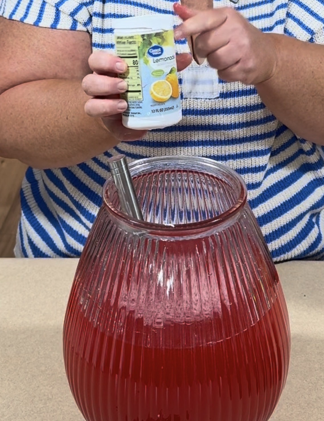 A person in a blue-and-white striped shirt adds a can of lemon concentrate to a large ribbed glass pitcher filled with a red drink. A mixing utensil is also inside the pitcher.