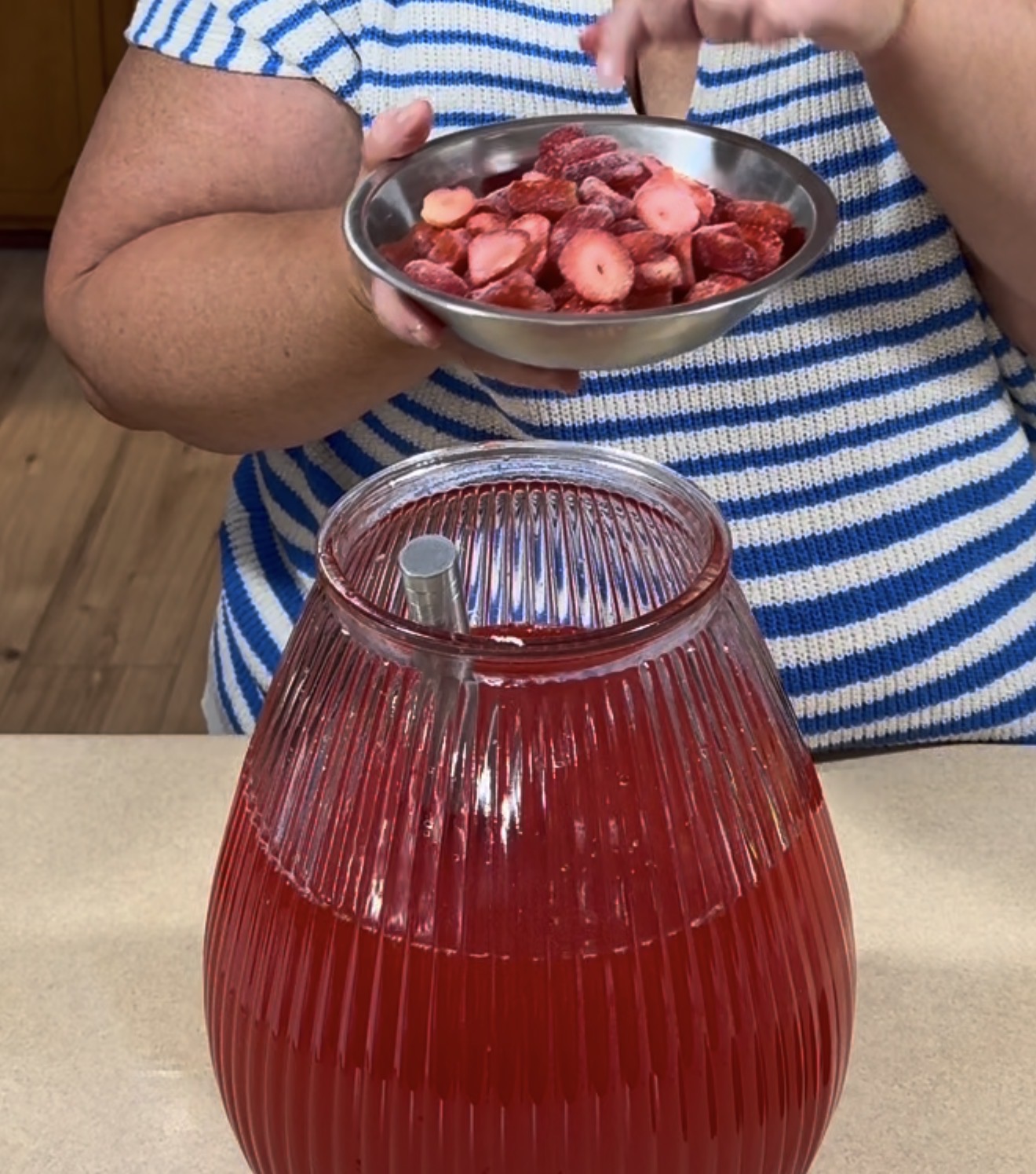A person in a striped shirt holds a small plate of sliced strawberries above a large glass jar filled with red juice and a metal straw on a countertop.