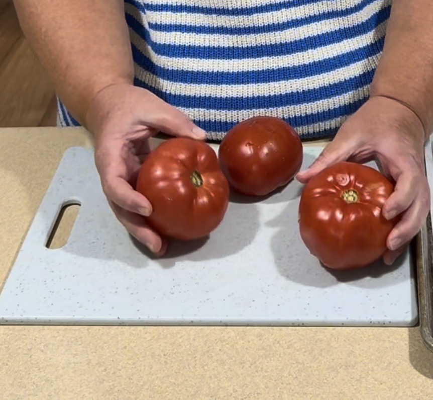 A person holding two red tomatoes over a white cutting board, with a third tomato resting on the board. The person is wearing a blue and white striped shirt.