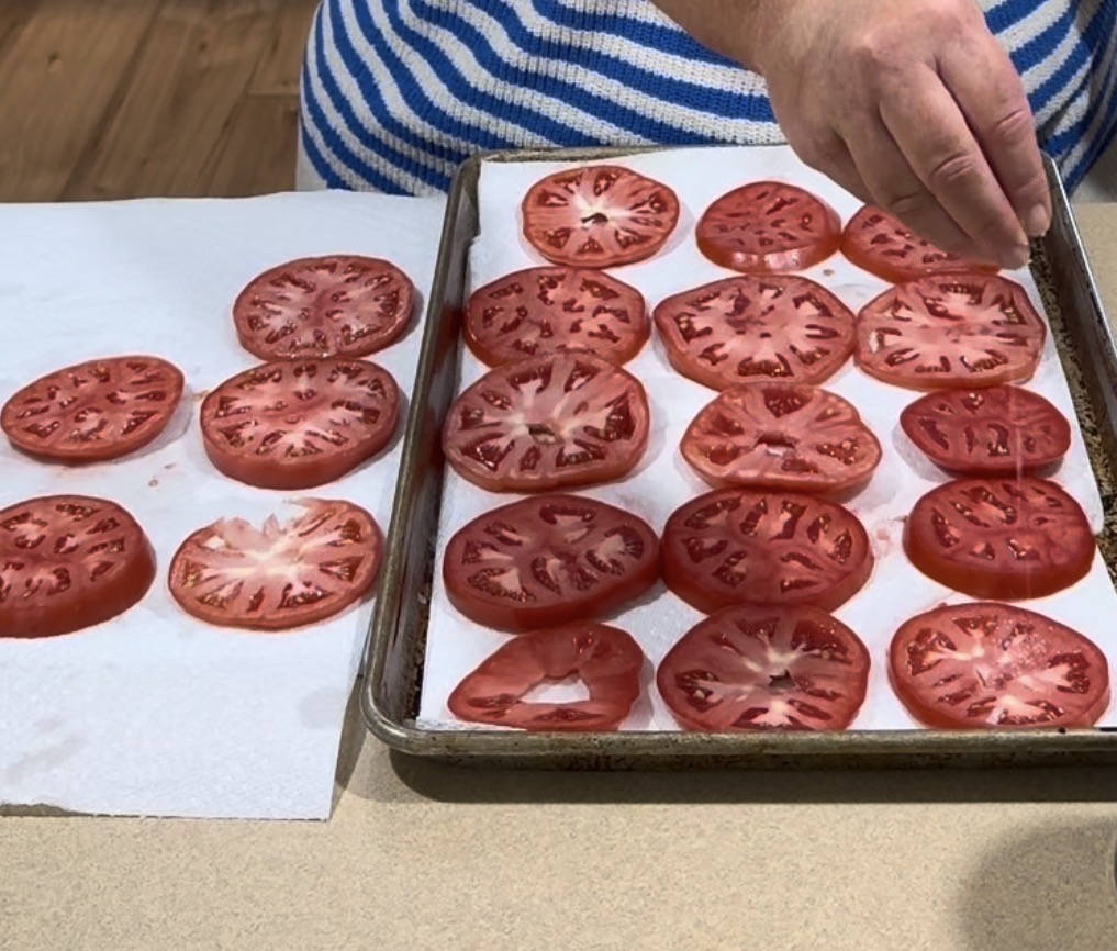 A person sprinkles salt on tomato slices arranged on a baking sheet lined with paper towels. More tomato slices sit on another paper towel next to the sheet.