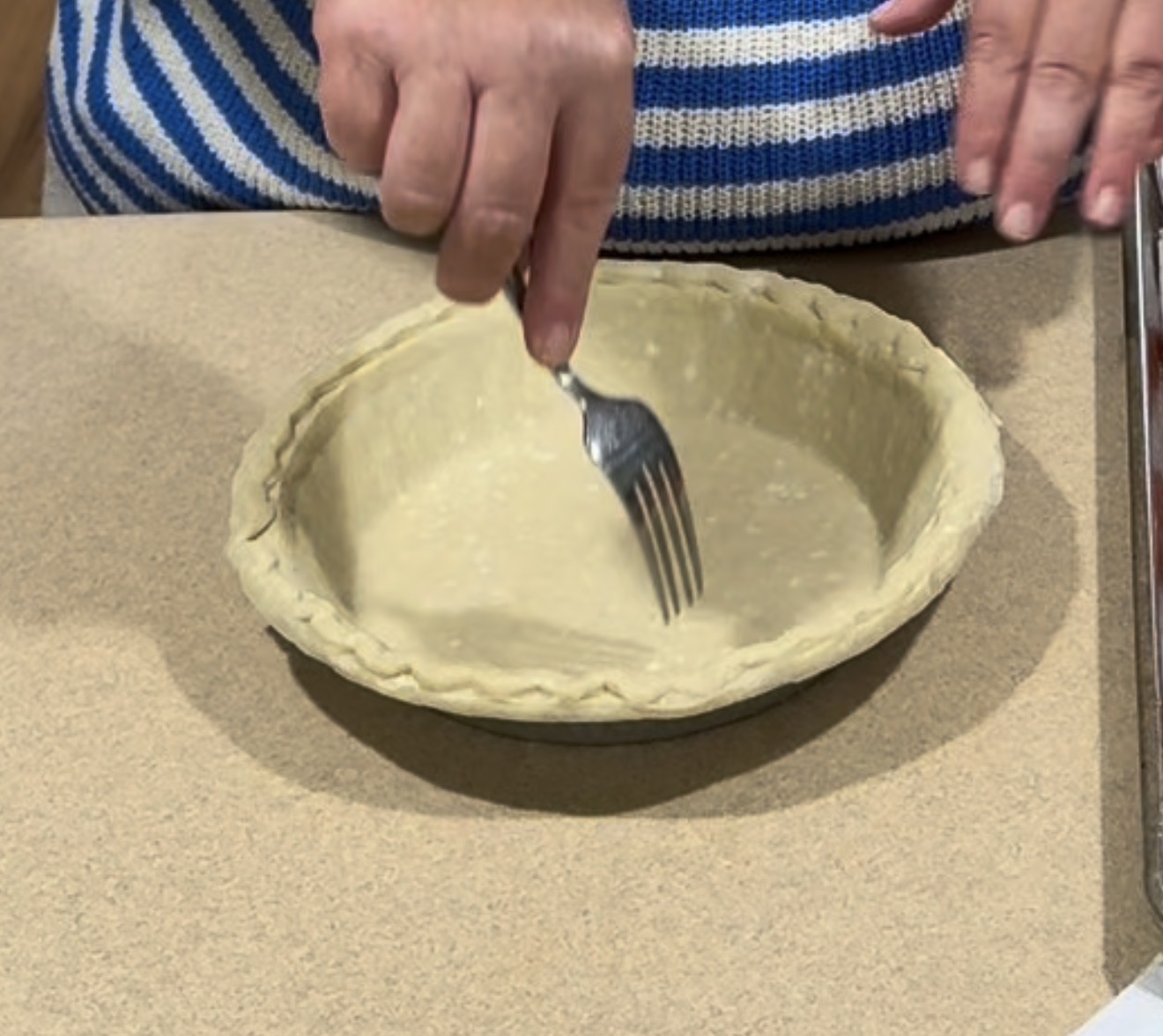 A person uses a fork to poke holes in an unbaked pie crust placed in a metal pie pan on a countertop.