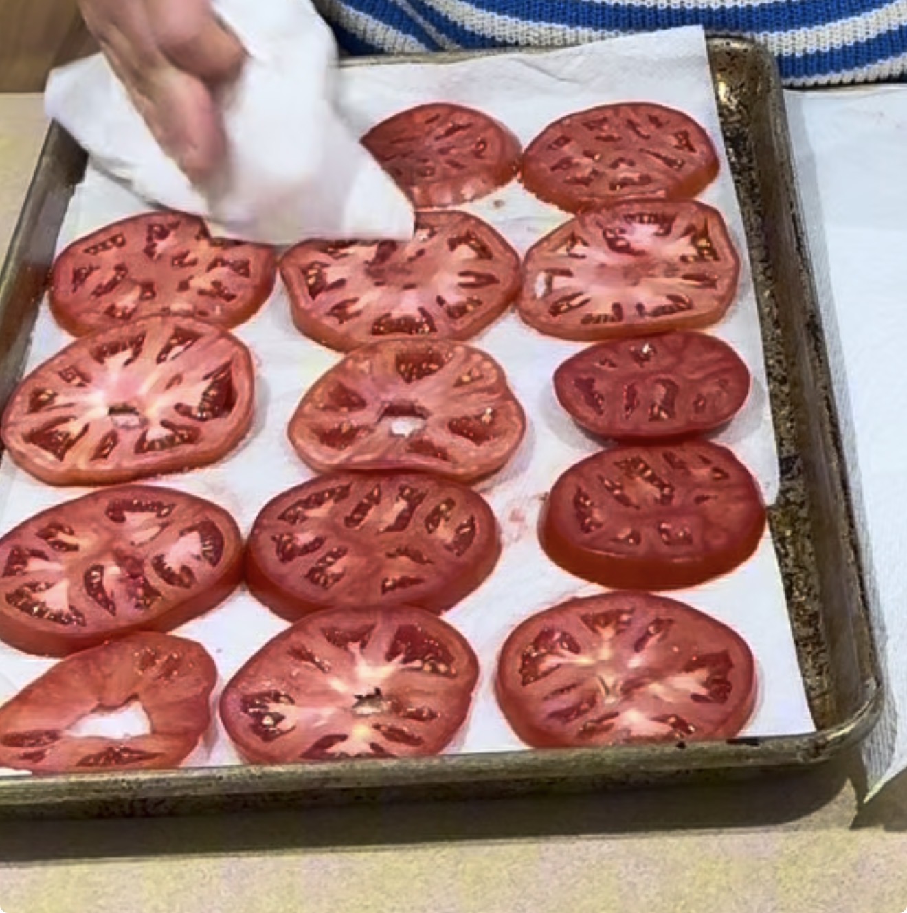 A hand uses a paper towel to blot moisture from sliced tomatoes arranged on a paper towel-lined baking sheet.
