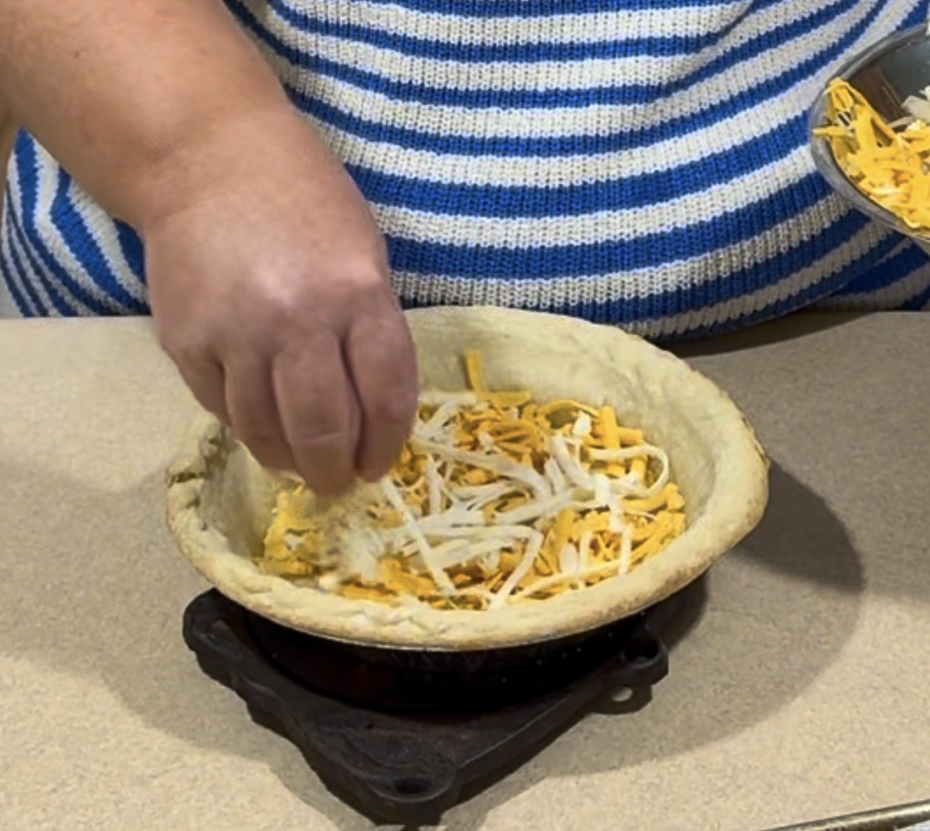 A person sprinkles shredded cheese into a partially filled pie crust, preparing a dish on a beige countertop.