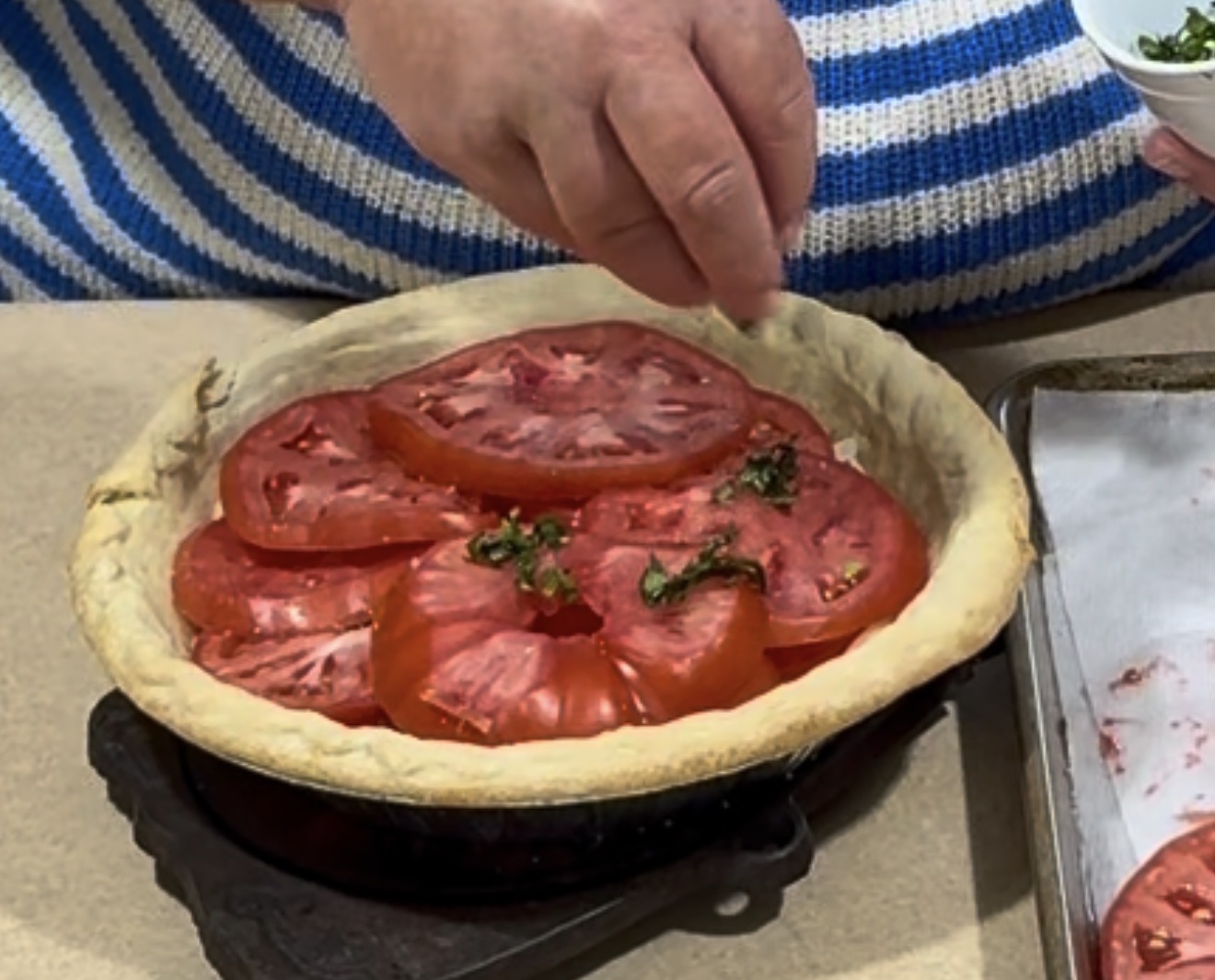 A hand sprinkles herbs over sliced tomatoes arranged in a pie crust, with a person wearing a blue and white striped sweater visible in the background.