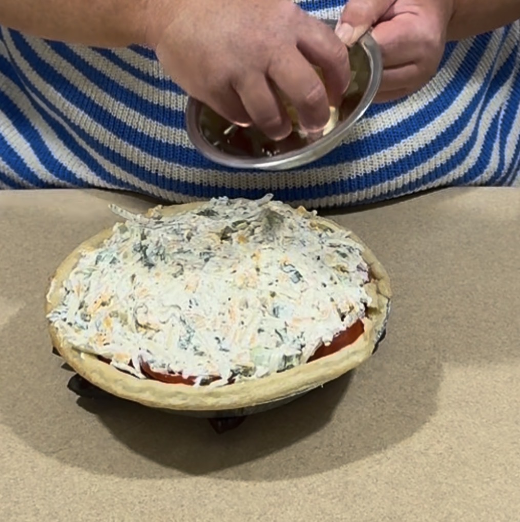 A person in a blue and white striped shirt adds ingredients from a small bowl to a pie topped with a creamy mixture on a countertop.