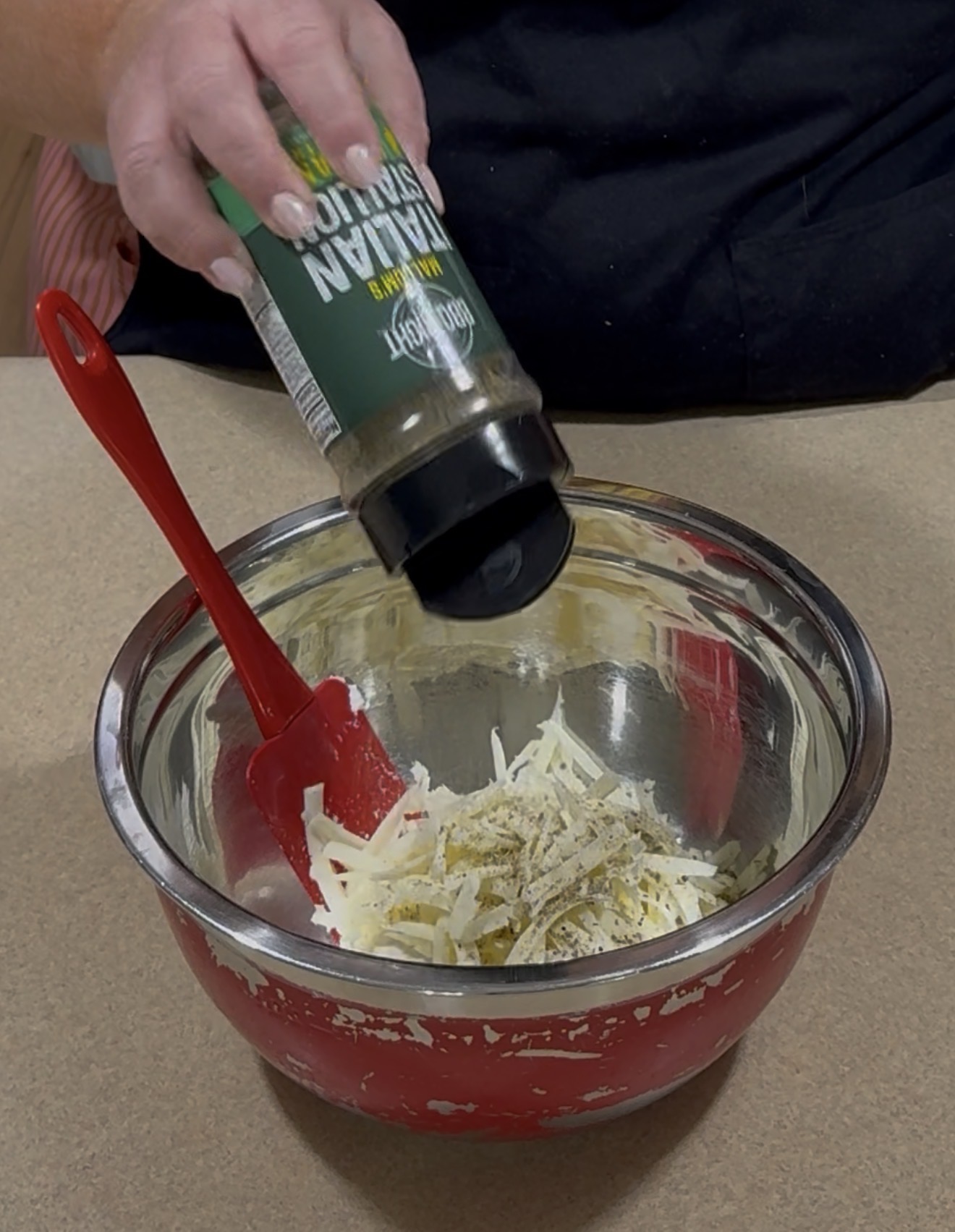 A hand sprinkles Italian seasoning into a metal mixing bowl filled with shredded cheese, using a red spatula to mix. The bowl sits on a beige countertop.