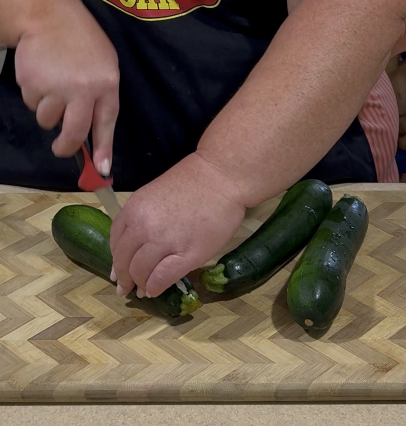 A person slices the end off a zucchini on a wooden cutting board, with two more zucchinis nearby.