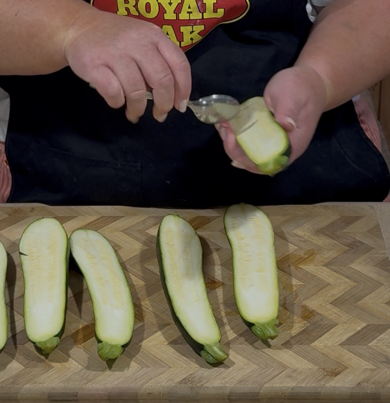 A person in a black apron scoops seeds from a halved zucchini with a spoon over a wooden cutting board with other zucchini halves.