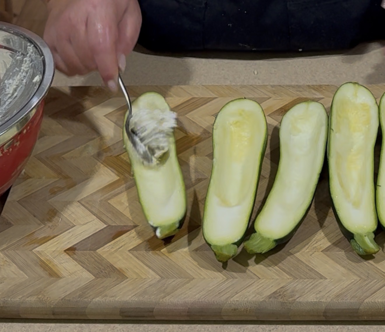 A hand uses a spoon to fill halved zucchini with a creamy mixture on a wooden cutting board, with a bowl of filling nearby.