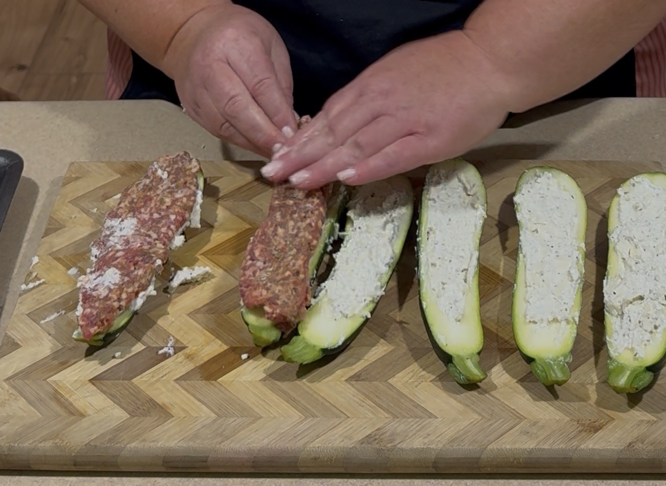 Hands are stuffing halved zucchinis with a mixture of ground meat and cheese on a wooden cutting board. Four zucchini halves are already filled with cheese.
